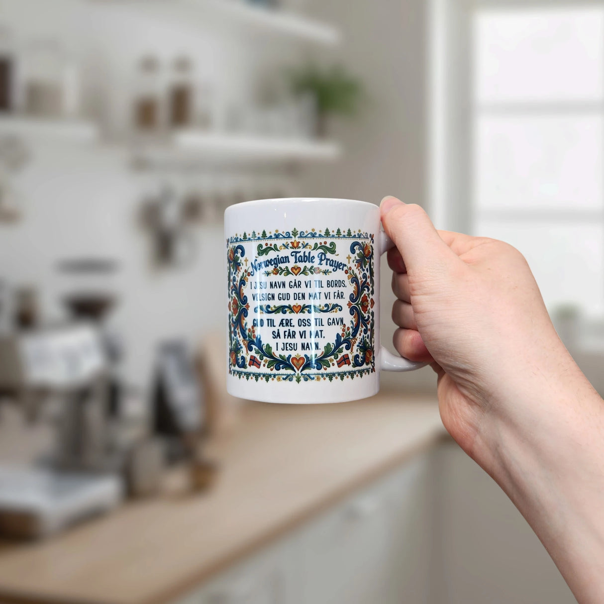 A person holds a white Nordic Wholesale Rosemaling mug with an intricate, colorful border and “Norwegian Table Prayer” text. In the background is a blurred kitchen with shelves, jars, and a coffee machine.