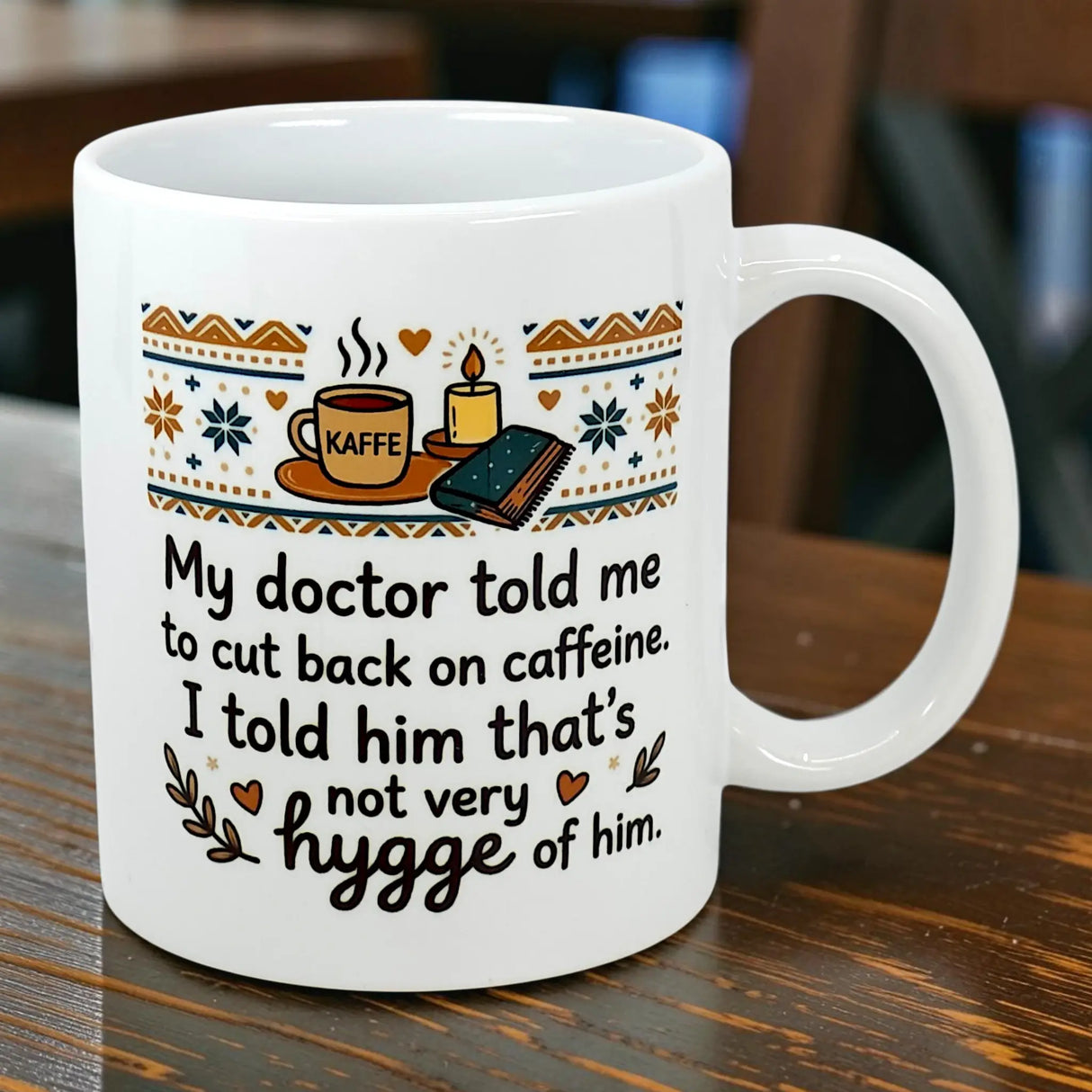 The Swedish Gift Store’s 11 oz “Not Very Hygge” Coffee Mug sits on a wooden table with coffee, a candle, and a book. Its text: My doctor told me to cut back on caffeine. I told him that’s not very hygge of him—true Scandinavian humor!.