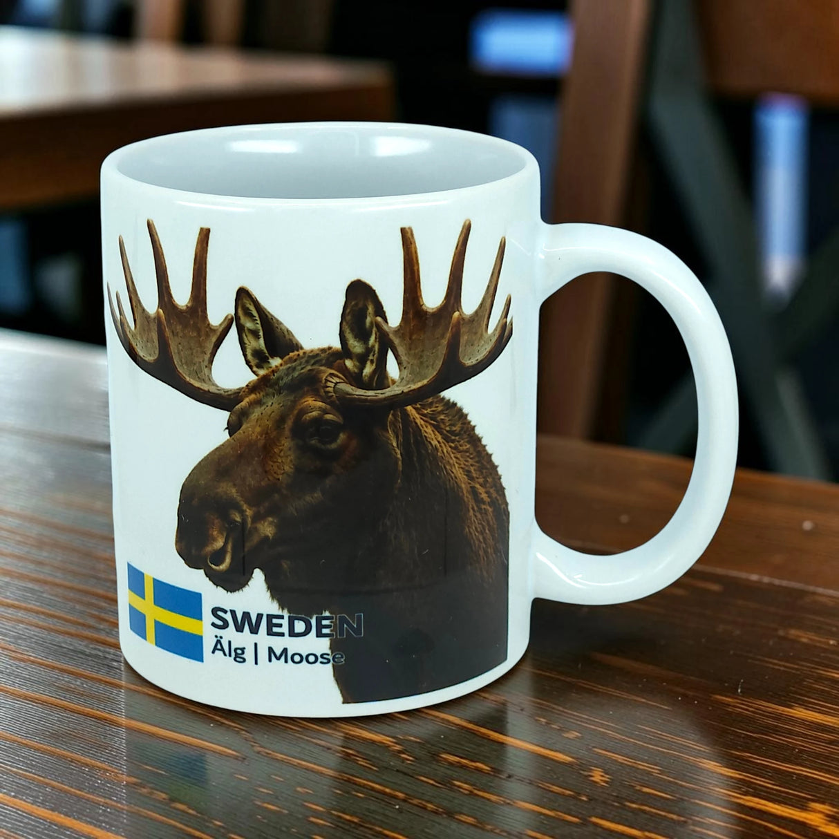 The Nordic Wholesale Mug: Sweden Älg Moose Ceramic Mug (11 oz) features a white ceramic design adorned with a photo of a moose, the Swedish flag, and the text "SWEDEN Älg | Moose." Placed on a wooden table, this charming mug is the perfect addition to any Scandinavian kitchen and beautifully celebrates Nordic nature.