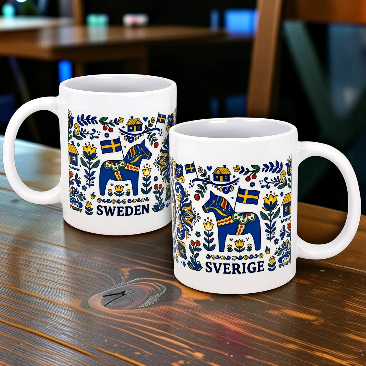 Two Nordic Wholesalers "Swedish Icons & Dala Horse Folk Art" ceramic mugs (11 oz) with colorful Dala horses, flowers, Swedish flags, and SWEDEN/SVERIGE text are displayed on a wooden table in a cozy indoor setting.