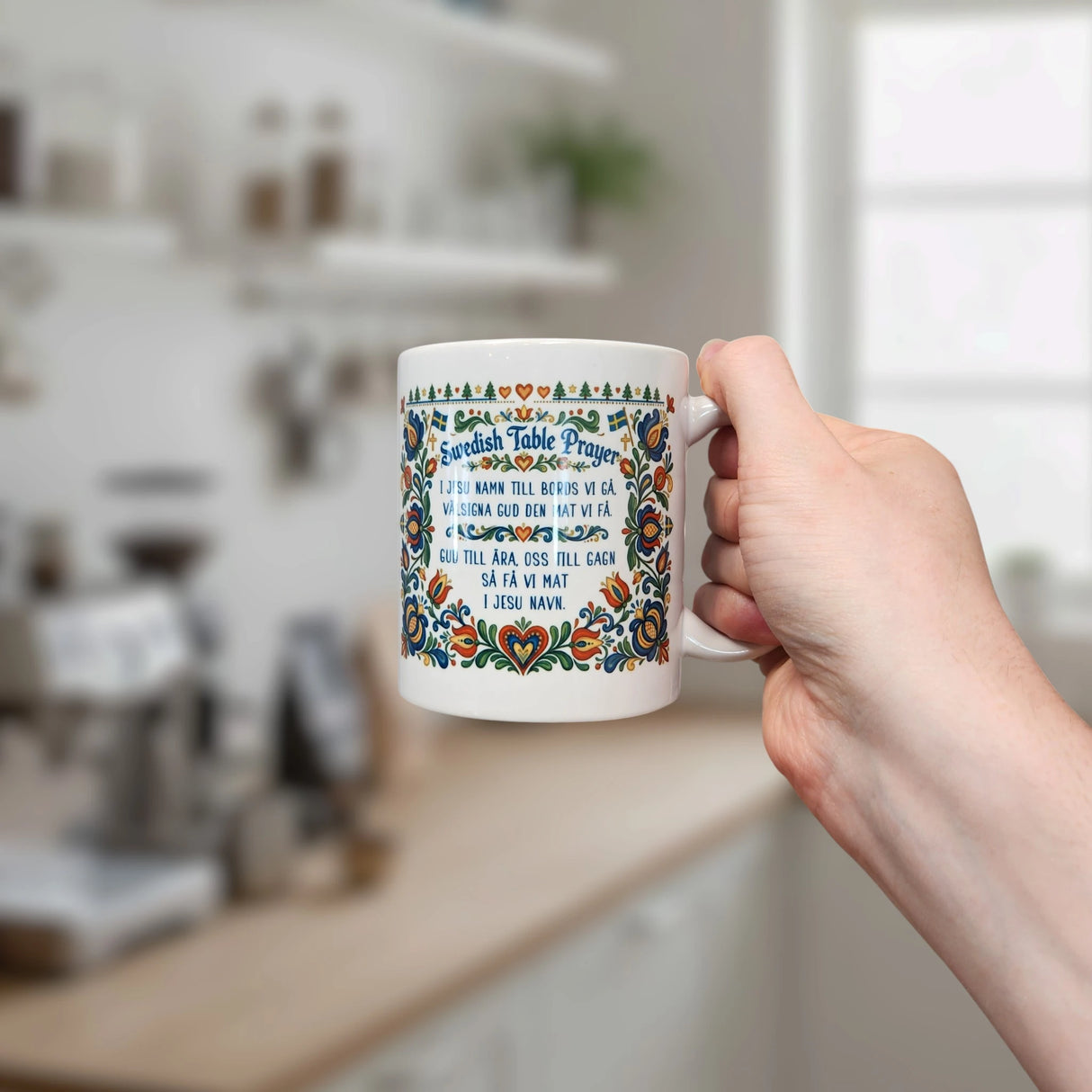 A hand holds the Nordic Wholesale Mug: Swedish Table Prayer Ceramic Kurbits Mug (11 oz Capacity) in a kitchen, with blurred shelves and appliances in the background.