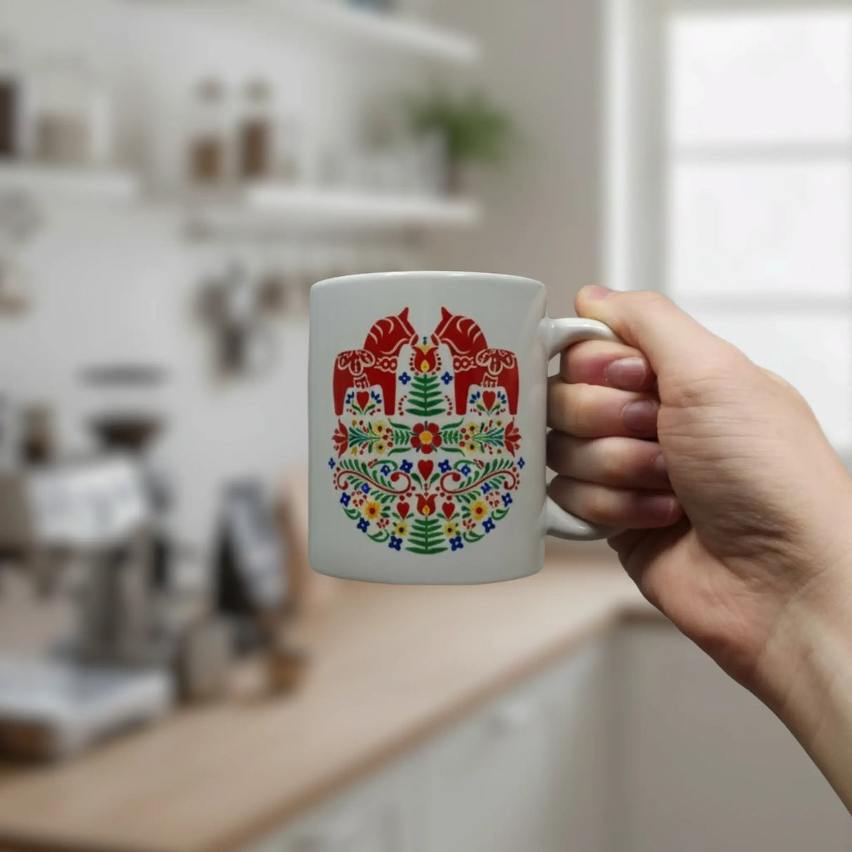 A hand holds the Swedish Gift Store’s 11oz ceramic mug, “Two Red Dala Horses in a Field of Swedish Folk Flowers,” featuring a colorful, symmetrical folk art pattern with red horses and florals; a kitchen background is visible.