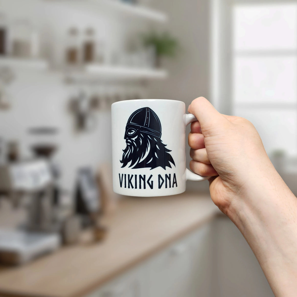 A hand holds the 11 oz Mug: Viking DNA White Ceramic Mug with Historically Accurate Helmet by Nordic Wholesale, featuring a black illustration of a Viking helmet and beard alongside the text "VIKING DNA," in a blurred kitchen setting.