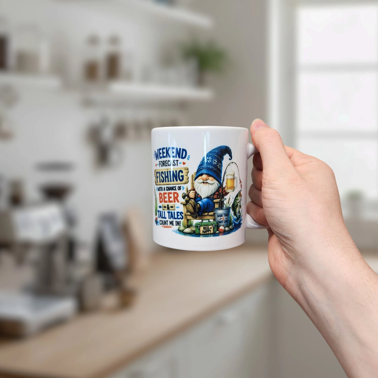A hand holds the 11 oz Nordic Wholesale Weekend Forecast Fishing Tomte Ceramic Mug in a kitchen, featuring a Scandinavian gnome and the text "Fishing with a chance of beer and tall tales"—an ideal fishing gift.