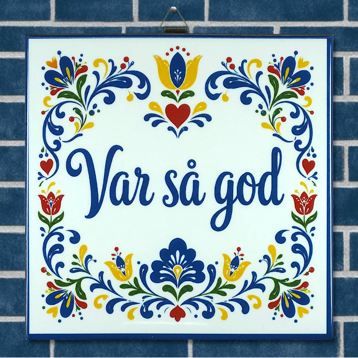 Ceramic trivet featuring blue floral patterns and the words “Var så god,” displayed on a blue tile background with Swedish folk art-inspired designs.