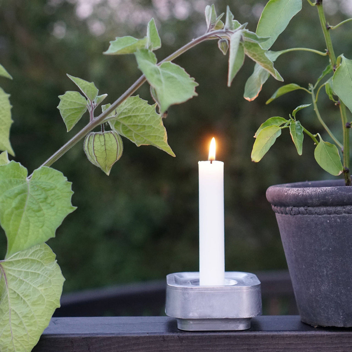 A white candle glows in the Stumpastaken Ettan holder, made from 100% recycled material (8.8x8.8x4 cm), placed on wood beside a potted plant and green foliage outdoors.