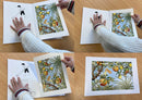 A person in a white sweater flips through a pop-up book featuring vintage illustrations of colorful birds and trees on a wooden table, drawing inspiration from the style seen in "Book: Art of Elsa Beskow 20- Pull-Out Frameable Prints.