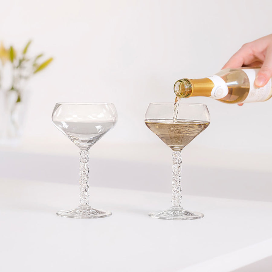 A hand elegantly pours wine into one of two Orrefors Carat Coupe 8.4oz glasses, featuring textured stems, beautifully arranged on a pristine white surface.