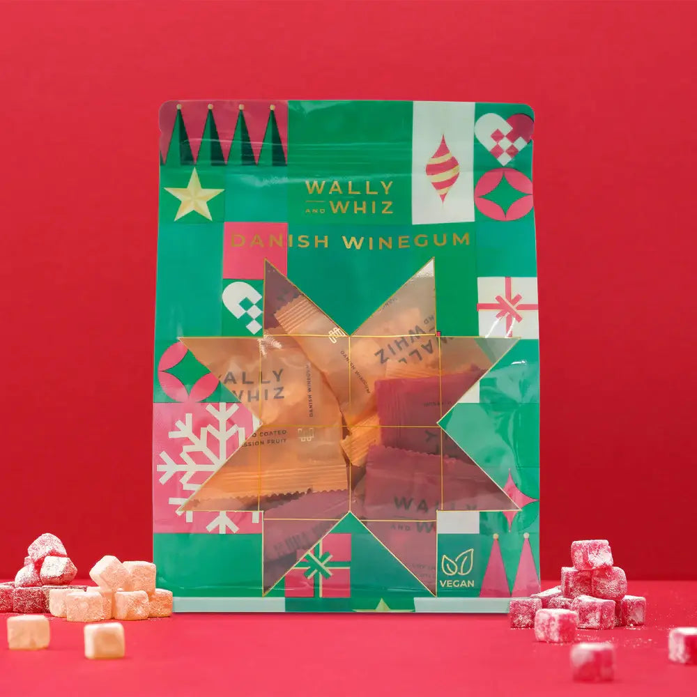 A festive Wally & Whiz Holiday Sampler Gift Bag 330g stands upright, its green and red packaging surrounded by assorted colorful vegan gummies on a red background—a cheerful treat for fans of Danish sweets.
