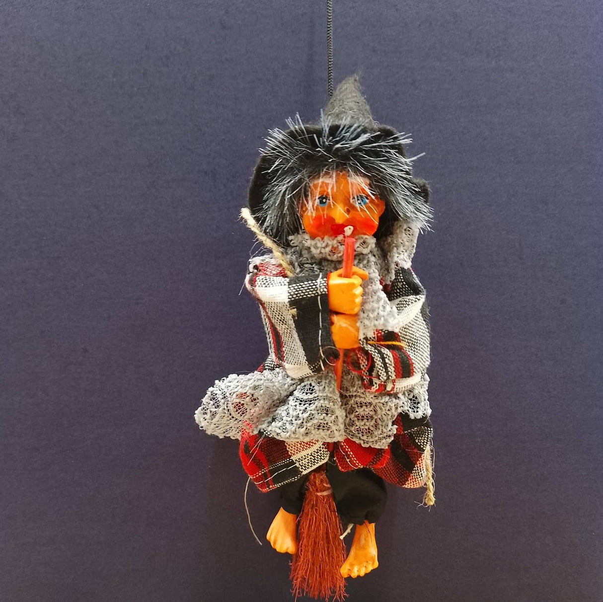 The Kitchen Witch: Minna Red Doll features a handmade orange face, hooded cloak, scarf, and holds a stick, displayed on a plain dark blue background—a charming piece of folklore décor.