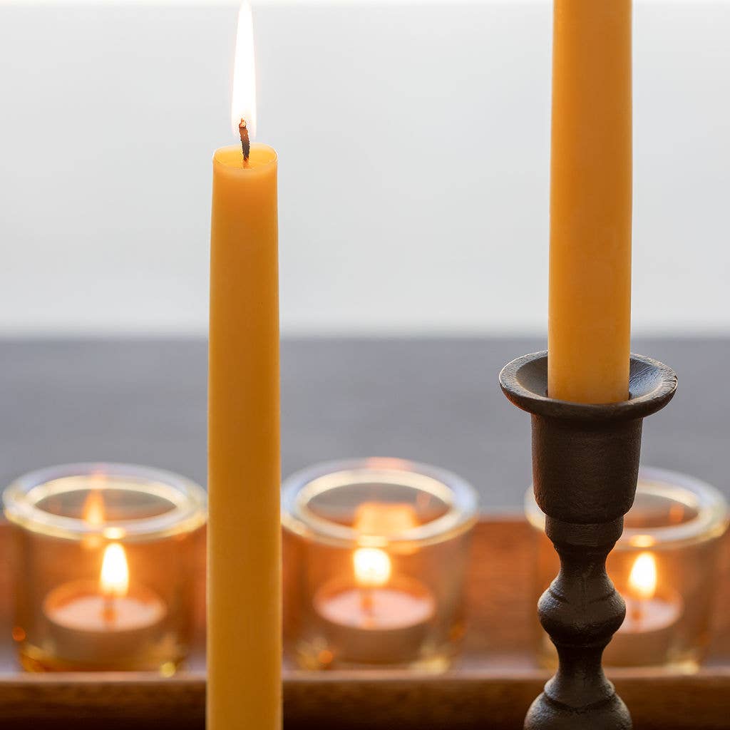 A lit yellow taper candle, made from pure beeswax, stands in a holder beside three Pure Beeswax Tea Light Candles – Handmade Raw/Clear Cup, nestled in glass holders on a tray.