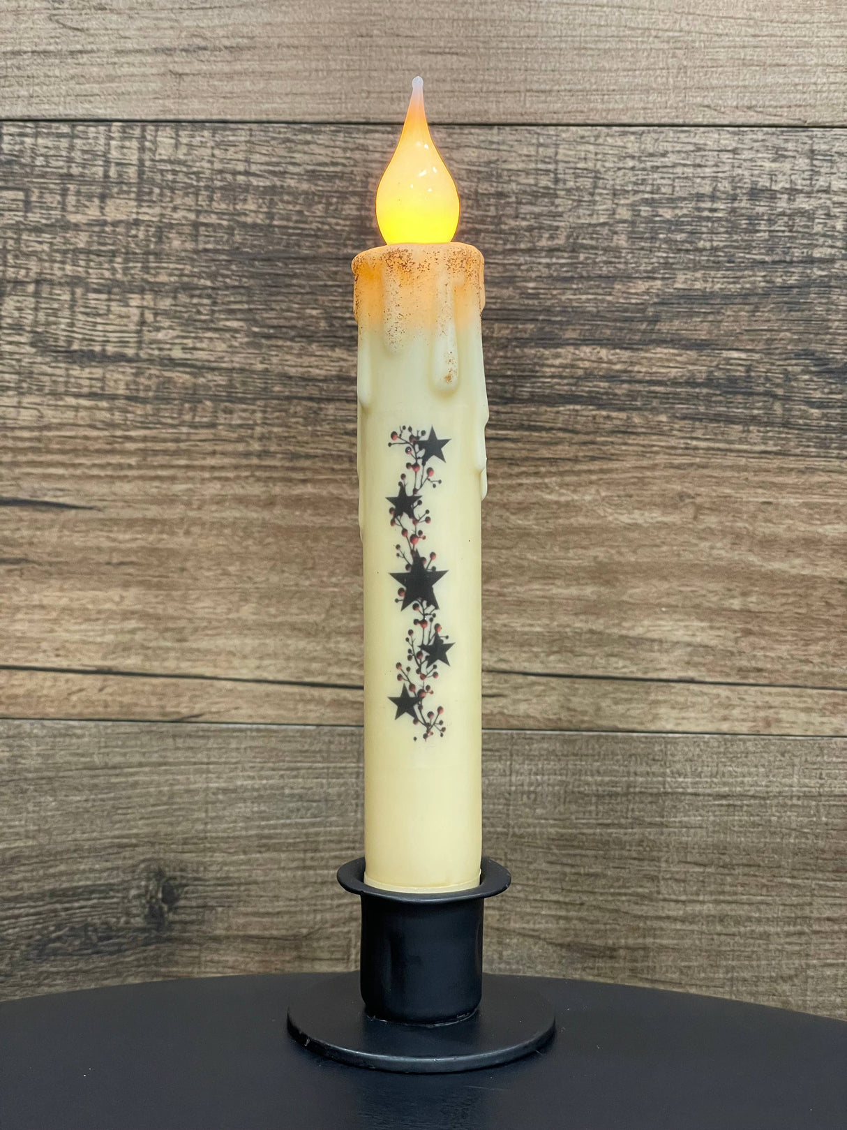A battery-operated candle featuring a melted wax look and star decorations is displayed in a Round Wrought Iron Taper Candle Holder, set against a wooden background.