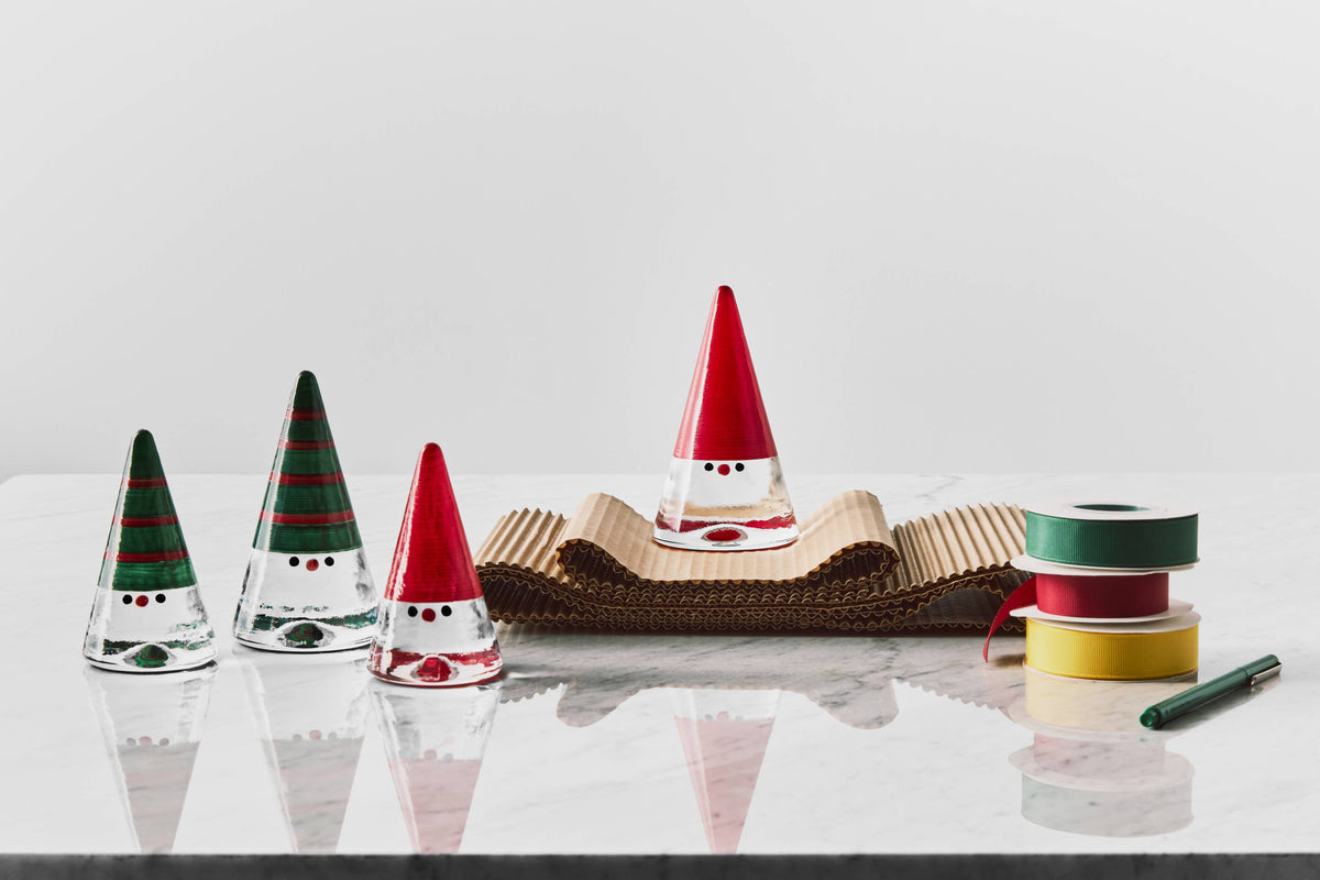 Four cone-shaped glass Tomte ornaments with hand-painted hats, arranged on a marble surface with rolls of ribbon.