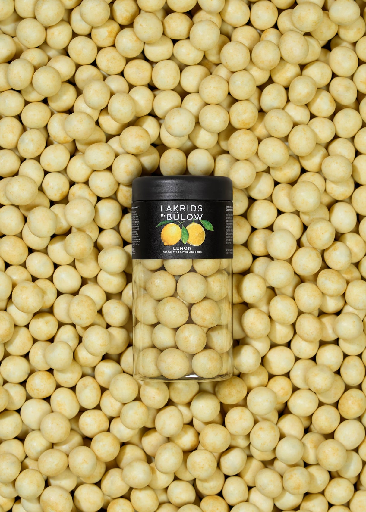 A jar of Lakrids by Bülow - Lemon - Danish Chocolate Coated Licorice (125g) is surrounded by vibrant yellow lemon chocolate licorice candies, highlighting this flavorful Danish treat.