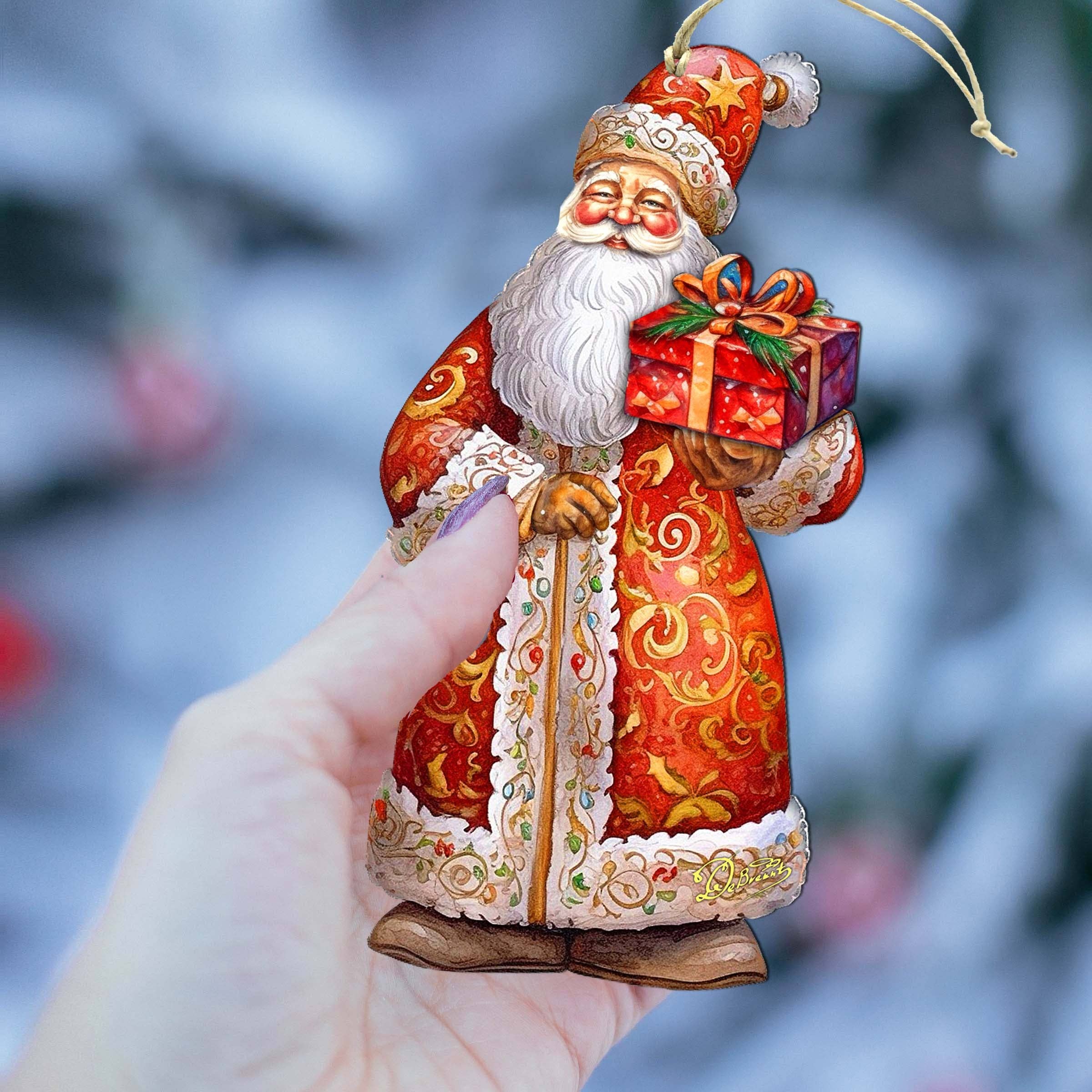 A hand holds the St. Nick's Gift Wood Ornament by G Debrekht, a 5.5" x 3" Christmas Santa dressed in red and gold, holding a wrapped gift, with a blurred winter background.