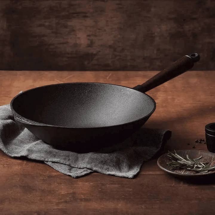 A Skeppshult Wok With Walnut Handle 12.2 / 30 cm sits on a cloth atop a wooden table, beside a plate of herbs and a small cup.