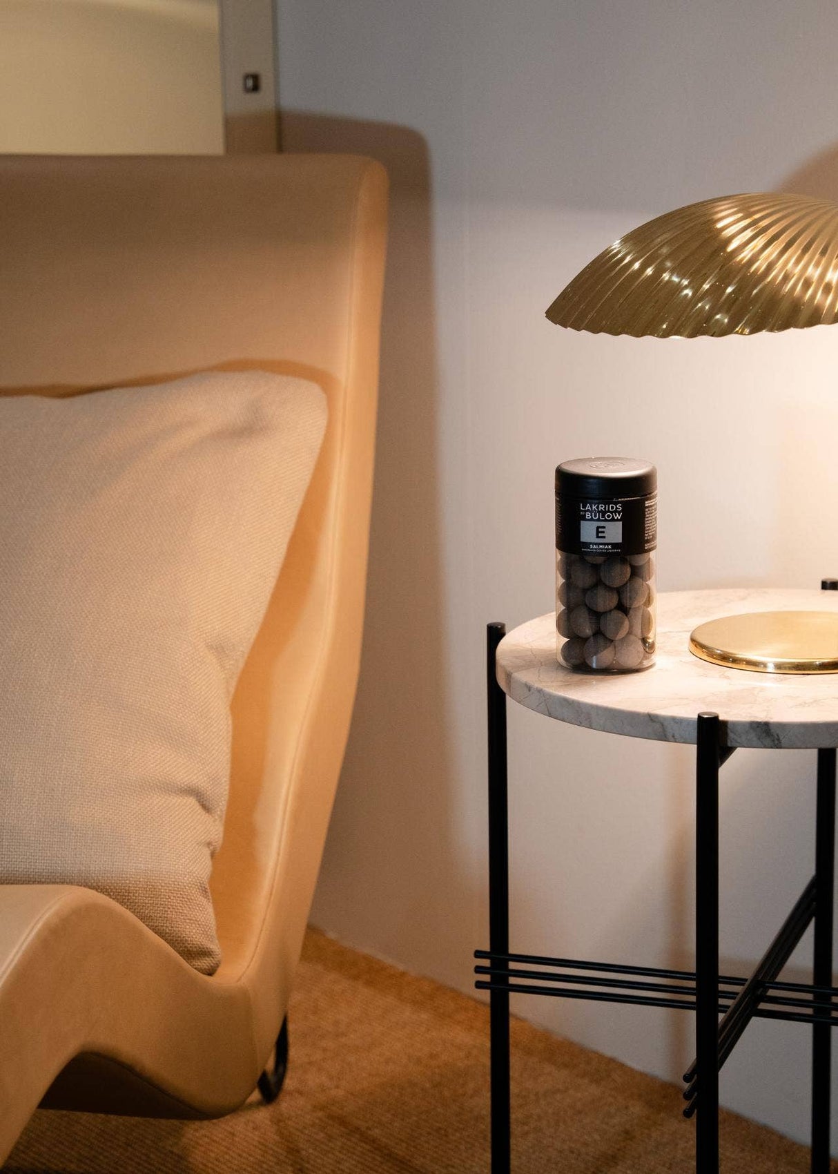 Beige chair with cushion beside marble-topped side table holding gold lamp, salty licorice jar, and capsules. Soft lighting, neutral interior.