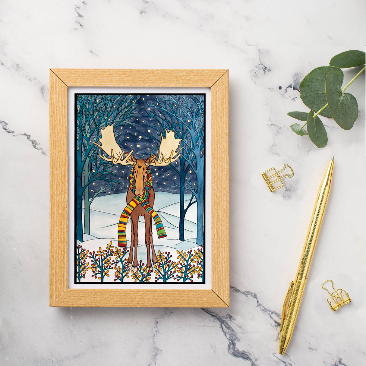 The Card: Holiday Moose by Sarah Angst is a 5x7 greeting card featuring a moose in a snowy forest with a striped scarf, blank inside, and comes with an envelope. Shown on marble with gold pen, clips, and green leaves.