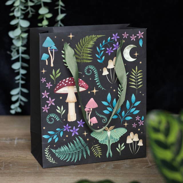 The Dark Forest Mushroom Bag is a black printed gift bag that artfully displays mystical flora and fauna through vibrant illustrations of mushrooms, ferns, leaves, flowers, and a crescent moon. It features green ribbon handles and rests gracefully on a wooden surface.