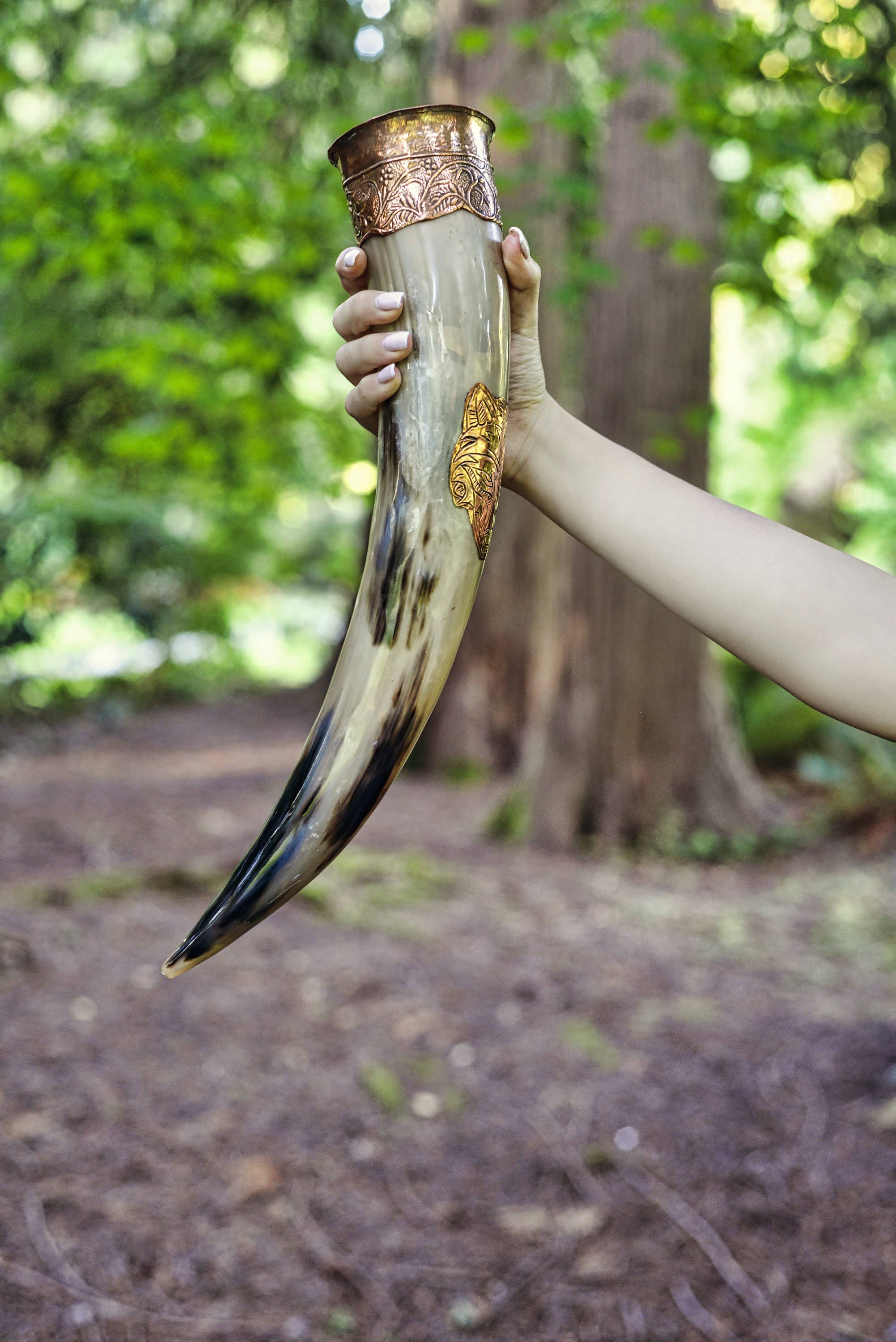 A person holds the Drinking Horn: Authentic Viking Wedding Gift, made from genuine ox horn and decorated with metal accents and intricate designs, set against a blurred forest background.