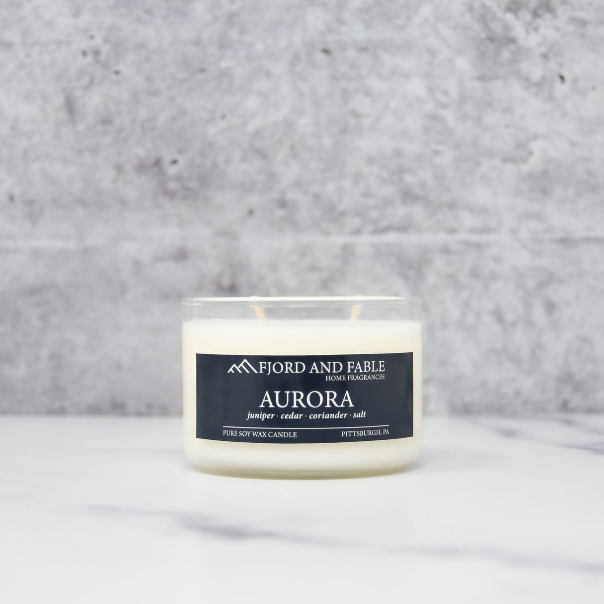 The Aurora Candle by Fjord & Fable is an 8oz Scandinavian-inspired soy wax candle in a clear jar, featuring notes of juniper, cedar, coriander, and salt, with a 45+ hour burn time and displayed elegantly on a marble surface.