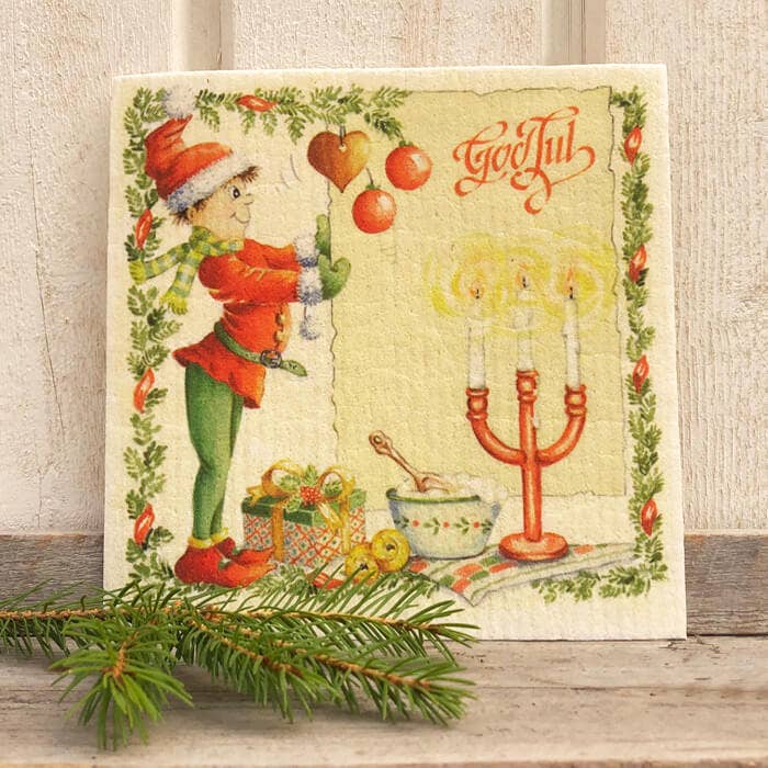 A festive card shows an elf hanging a heart ornament, gifts, and candles with "God Jul" text. Next to a pine sprig is the Straw Goat "Julbock" dish cloth featuring the classic Christmas goat with red ribbons.