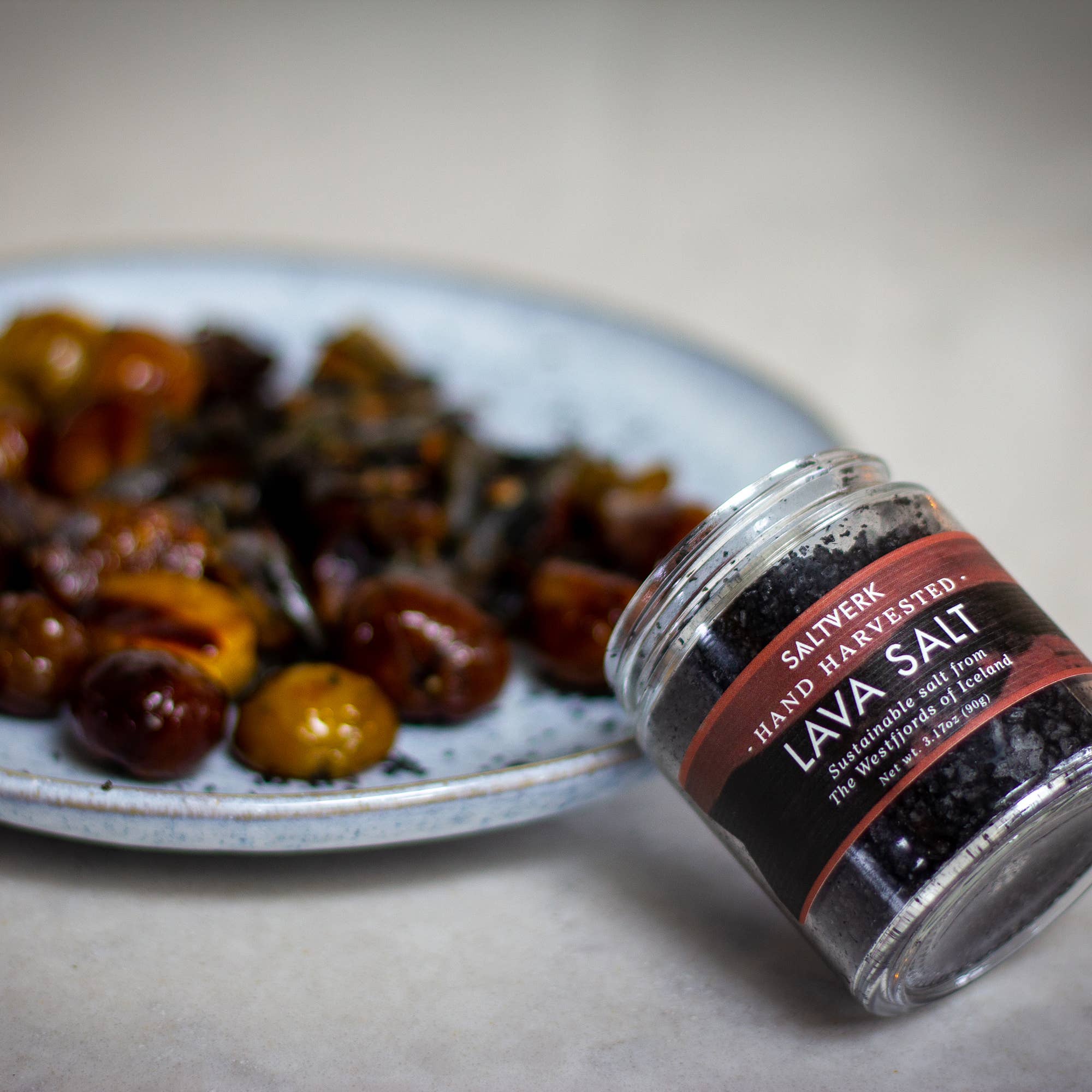 A jar from the SALTVERK Gift Set—featuring Flaky Sea Salt, Lava Salt, Birch Smoked Salt, and Arctic Thyme Salt—rests on its side beside a plate of roasted cherry tomatoes on a light surface.