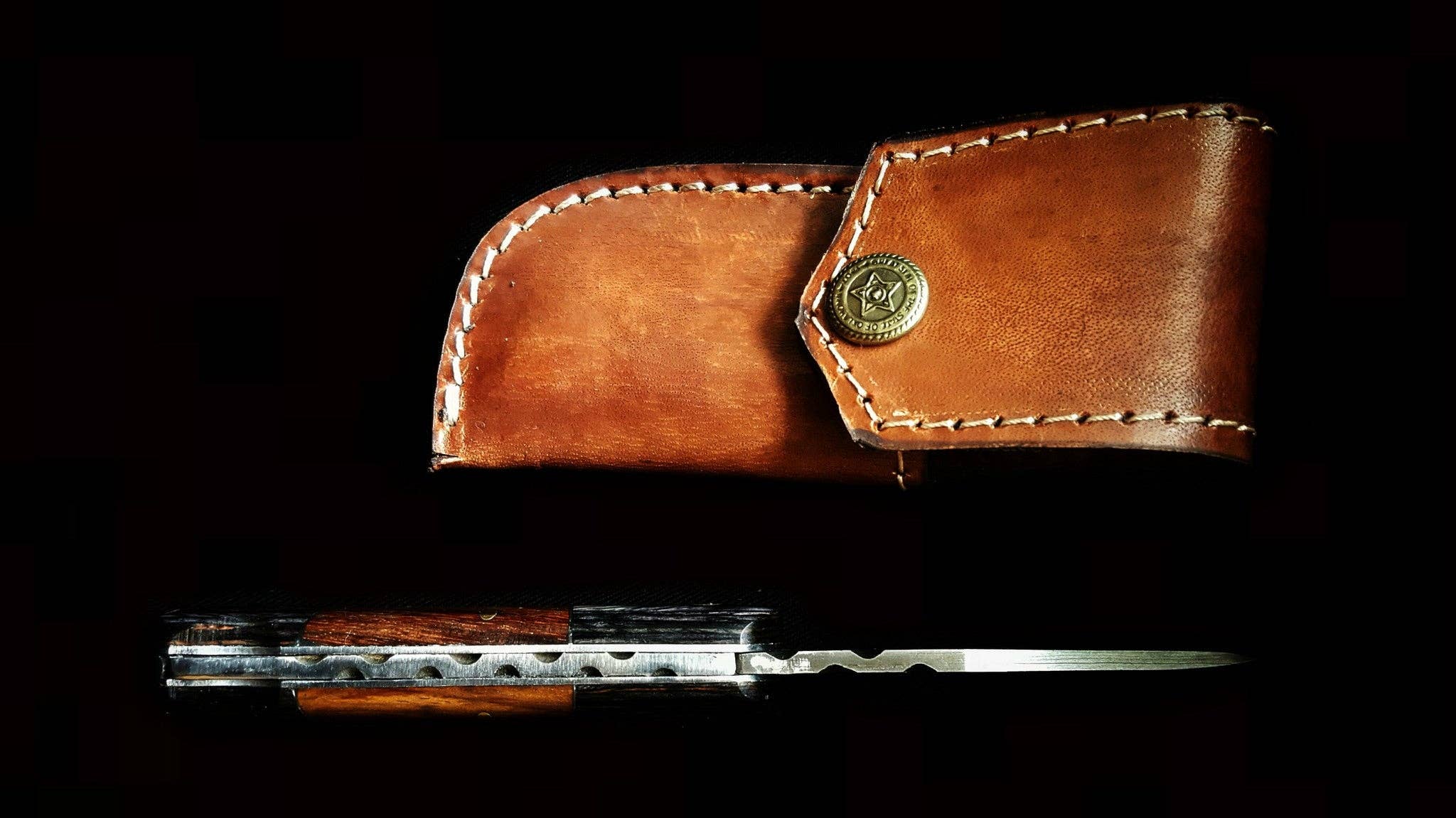 The Damascus Steel Pocket Knife features a patterned blade and walnut wood handle placed below its brown leather sheath with a brass button, all displayed against a black background.