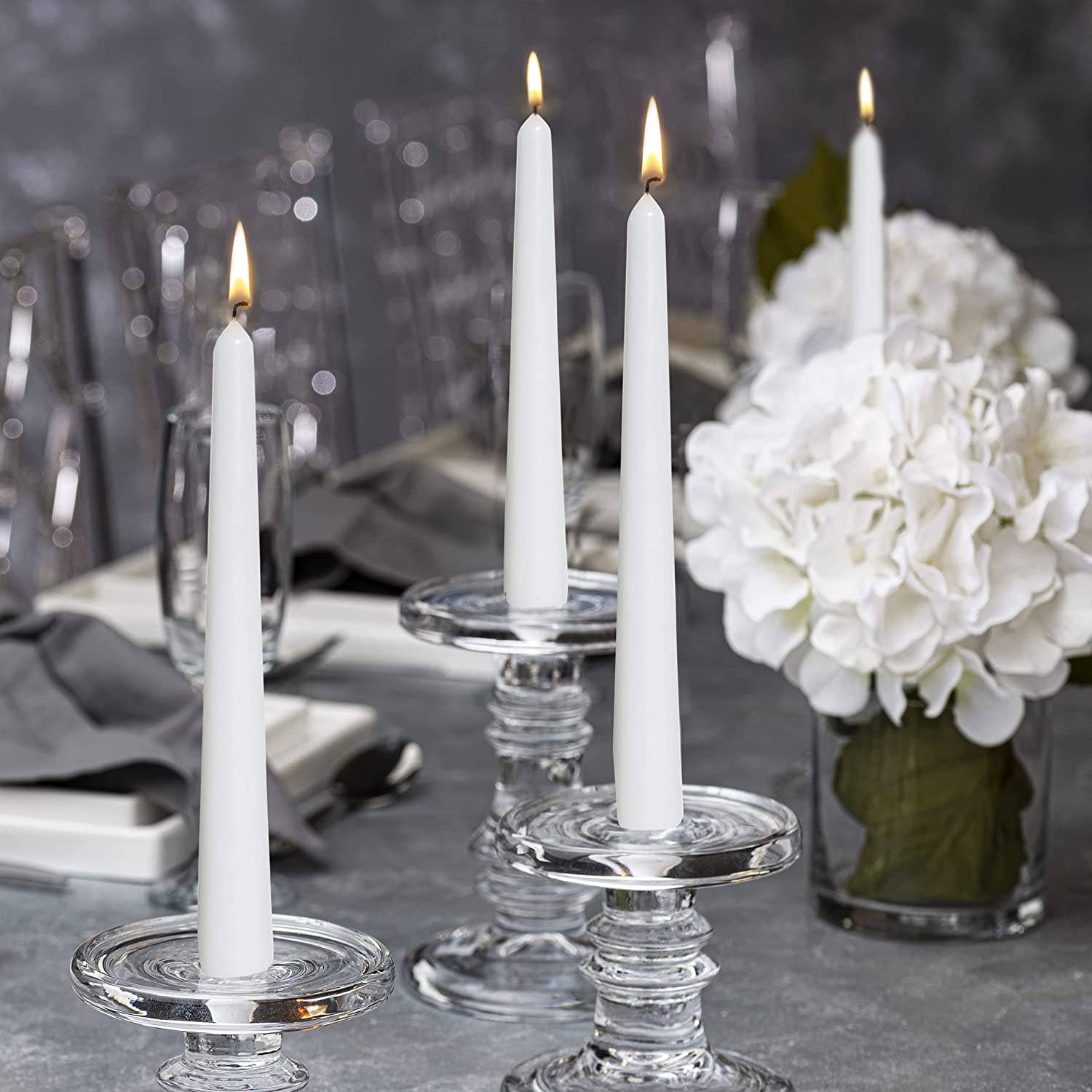 Four lit White 10 Inch Unscented Taper Candles (4 Pack) are arranged on a dining table with plates, gray napkins, clear glass holders, and white hydrangea centerpieces.