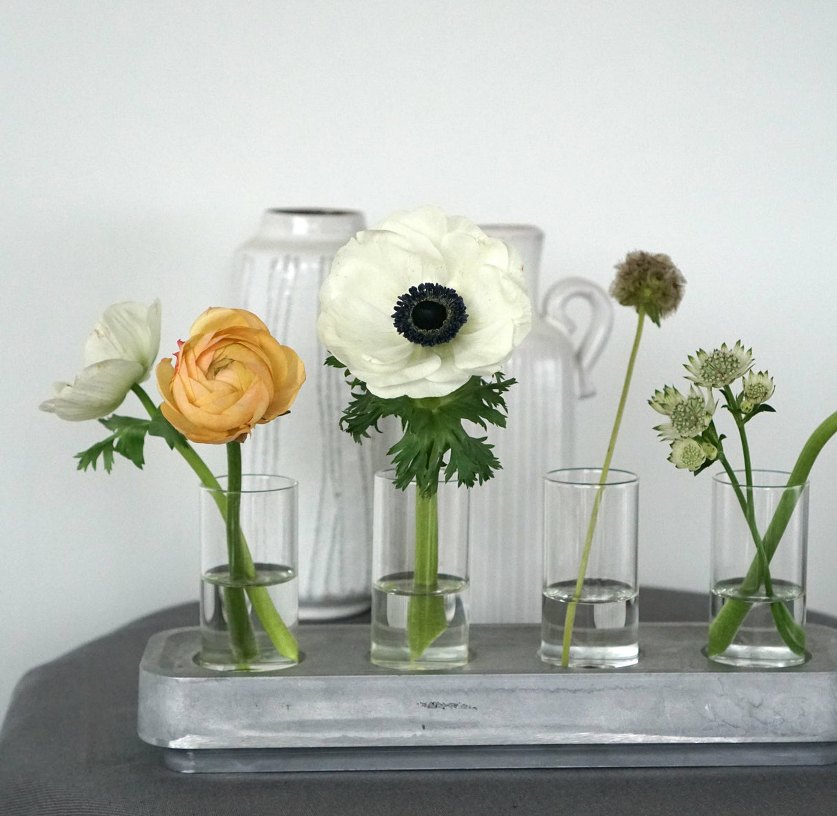 Four flowers in glass vases sit on a tray with varied colors and types, elegantly arranged against a gray and white ceramic background. They are complemented by the Stumpastaken Swedish Candle Holder crafted from recycled aluminum, highlighting eco-friendly beauty.