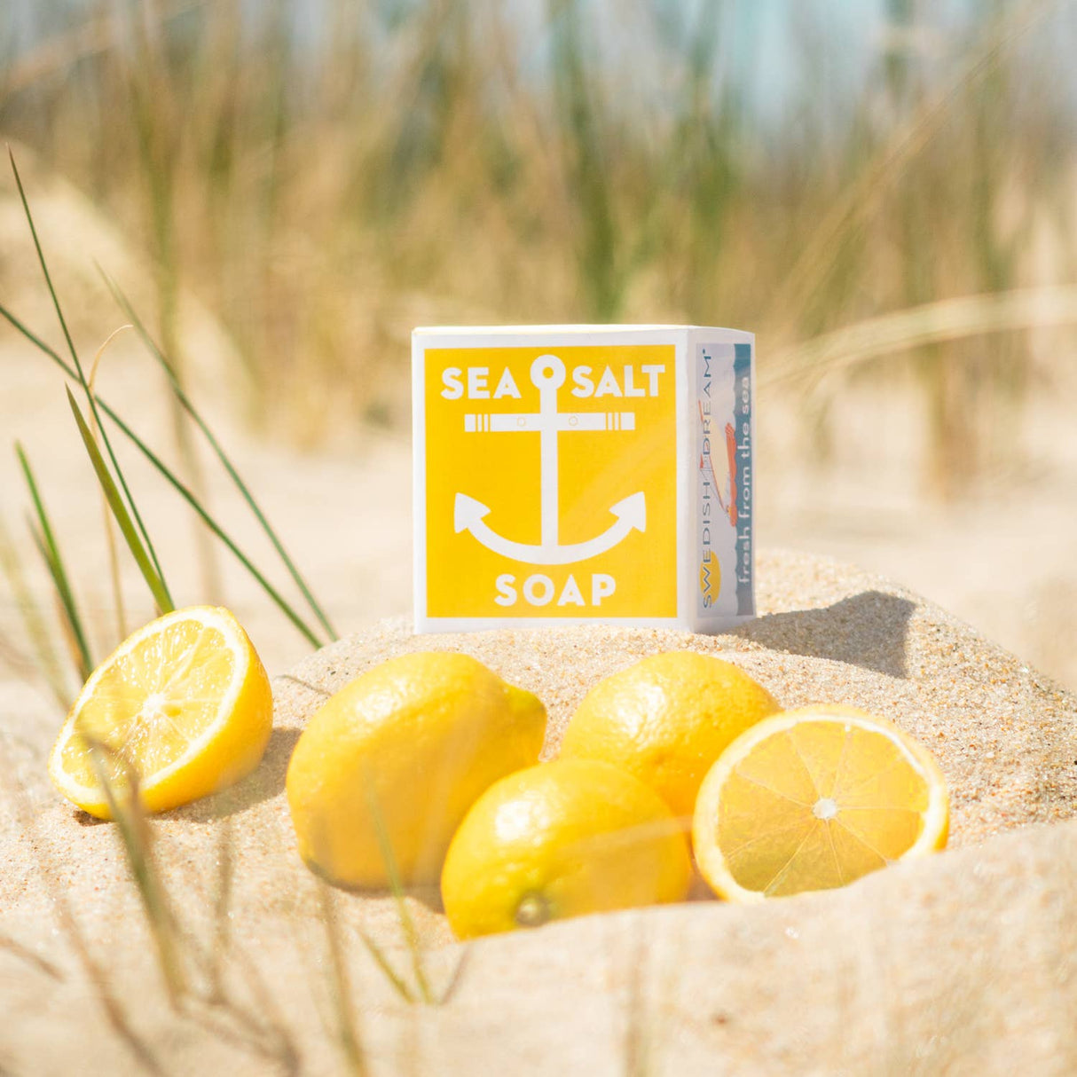 A 4 oz bar of Swedish Dream Sea Salt Lemon Soap featuring a yellow anchor design rests on sand beside whole and sliced lemons, with beach grass in the background.