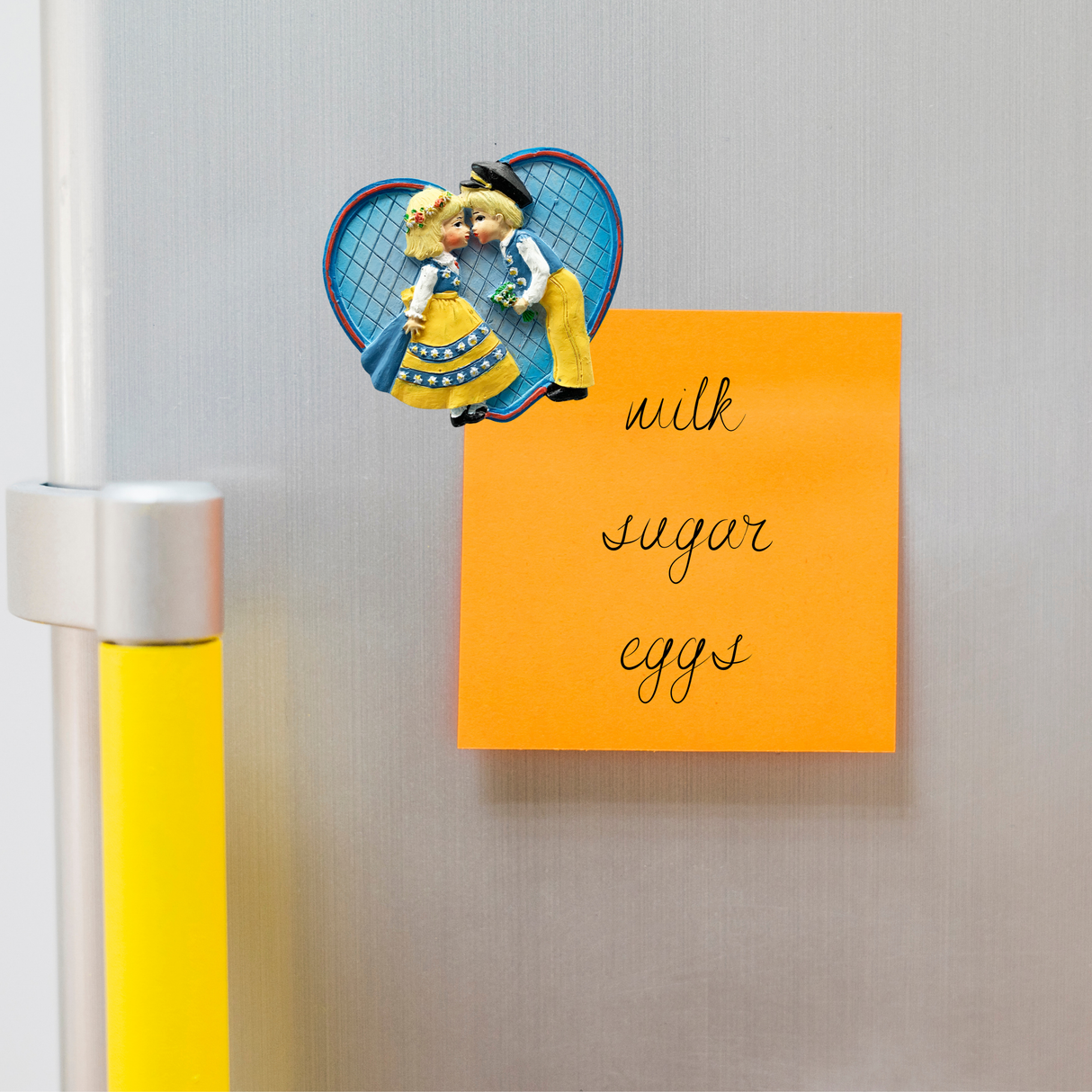 An eye-catching orange sticky note adorns the refrigerator door, outlining essentials such as milk, sugar, and eggs. Next to it, a charming Magnet: Swedish Kissing Couple Party Favor Kitchen Magnet in traditional attire turns an ordinary list into a delightful novelty gift display.