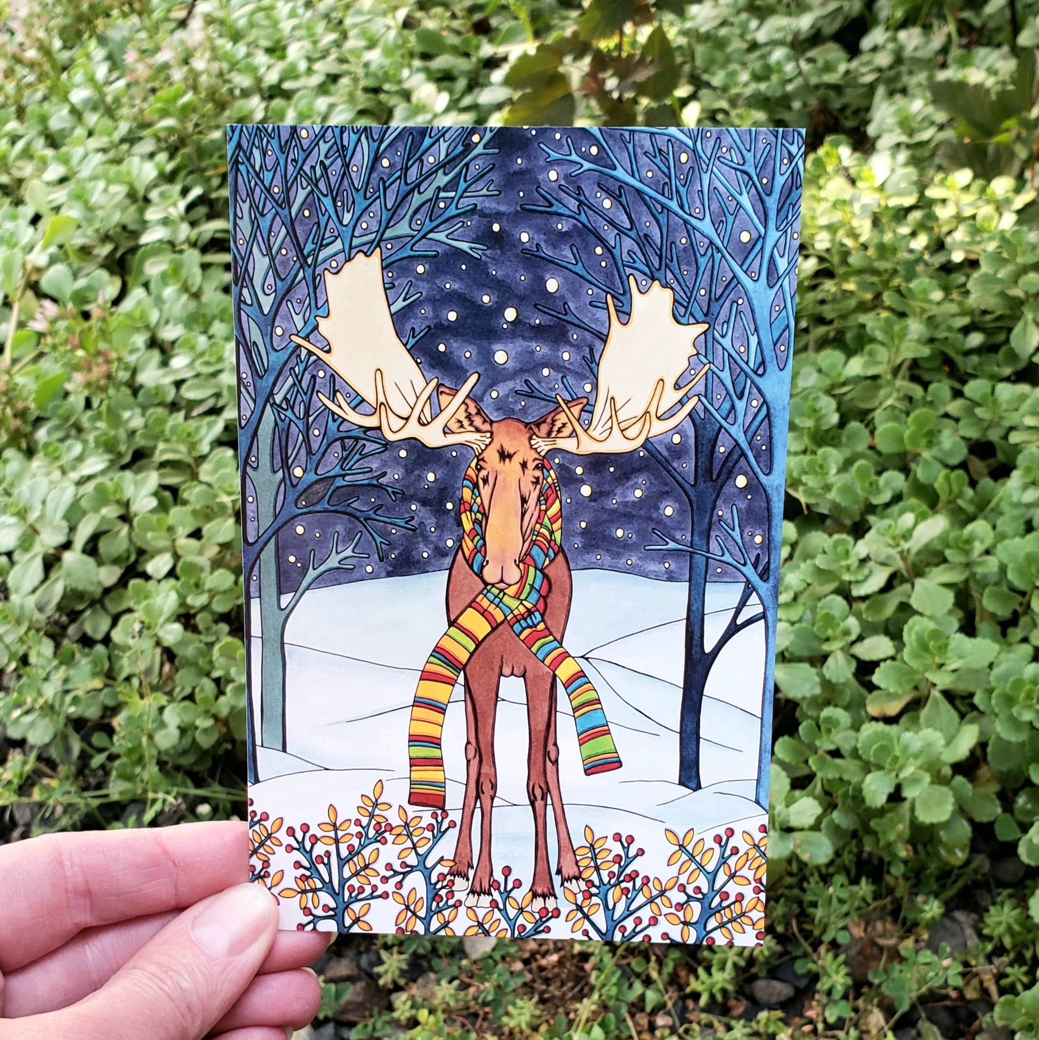 A hand displays the "Postcard: Holiday Moose by Sarah Angst 4x6 inches," showing a moose in a striped scarf in a snowy, starry forest with bare trees, all set against green outdoor foliage.