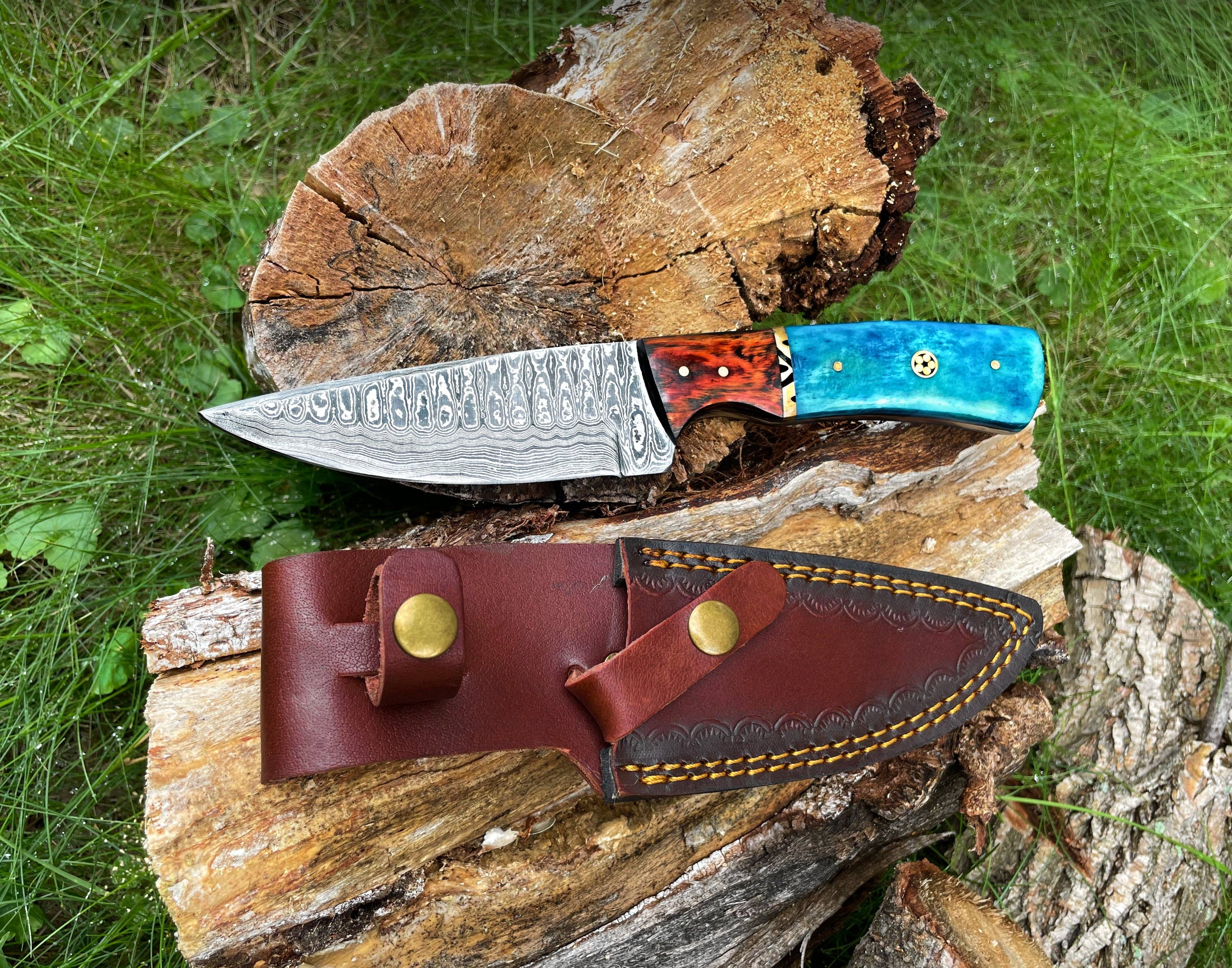 A Hand Forged Damascus Knife with a drop-style blade and black handle rests on a tree stump, its brown leather sheath below, surrounded by grass and wood in the background.