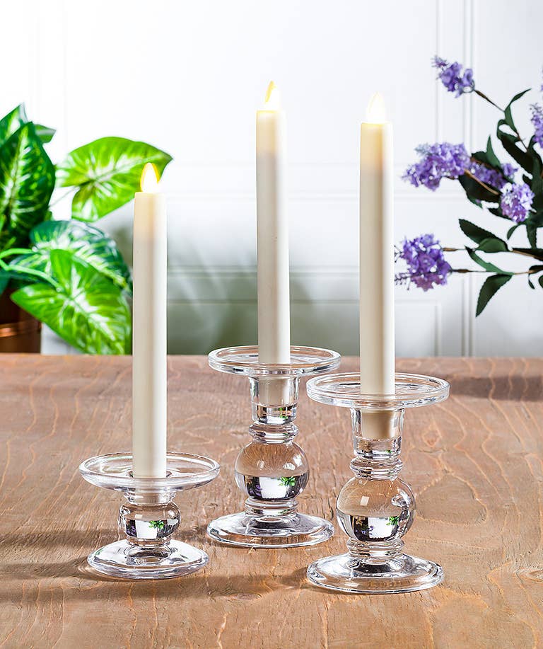Three lit white taper candles in Small Reversible Candle Holders (3.5"L) are arranged on a wooden table, with green and purple plants in the background.