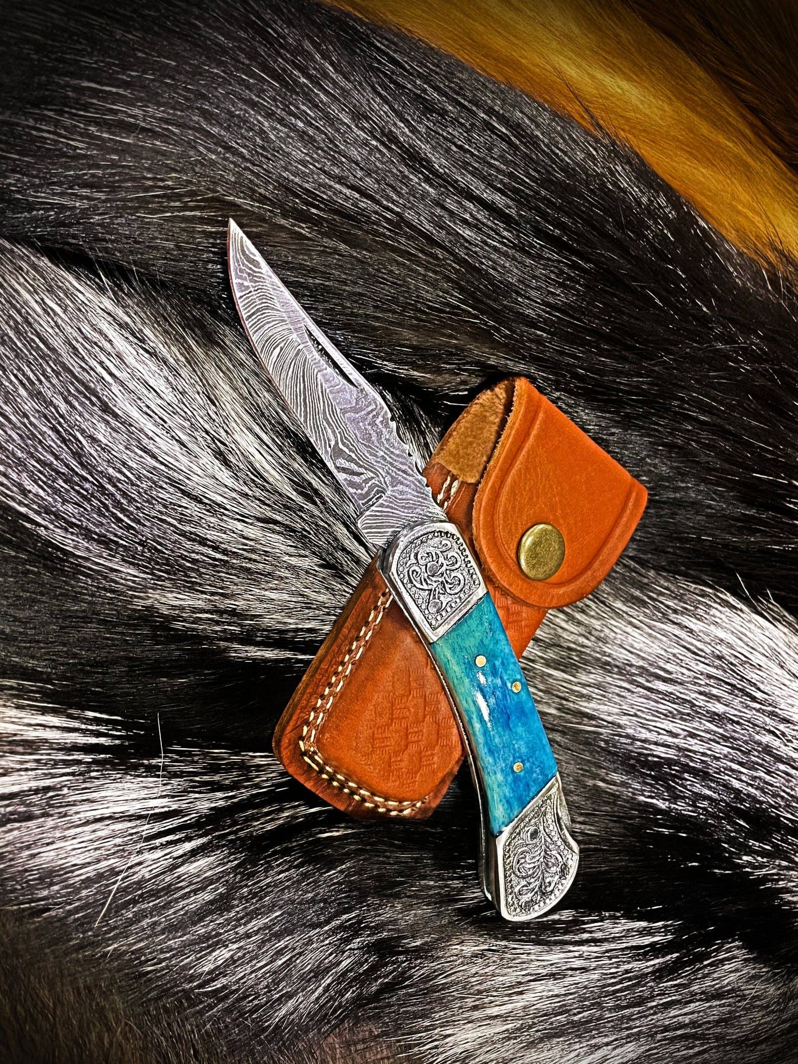 The Knife: Dyed Blue Bone 6.5 Damascus Steel features a patterned blade and blue bone handle, resting on a leather sheath, displayed atop animal fur.