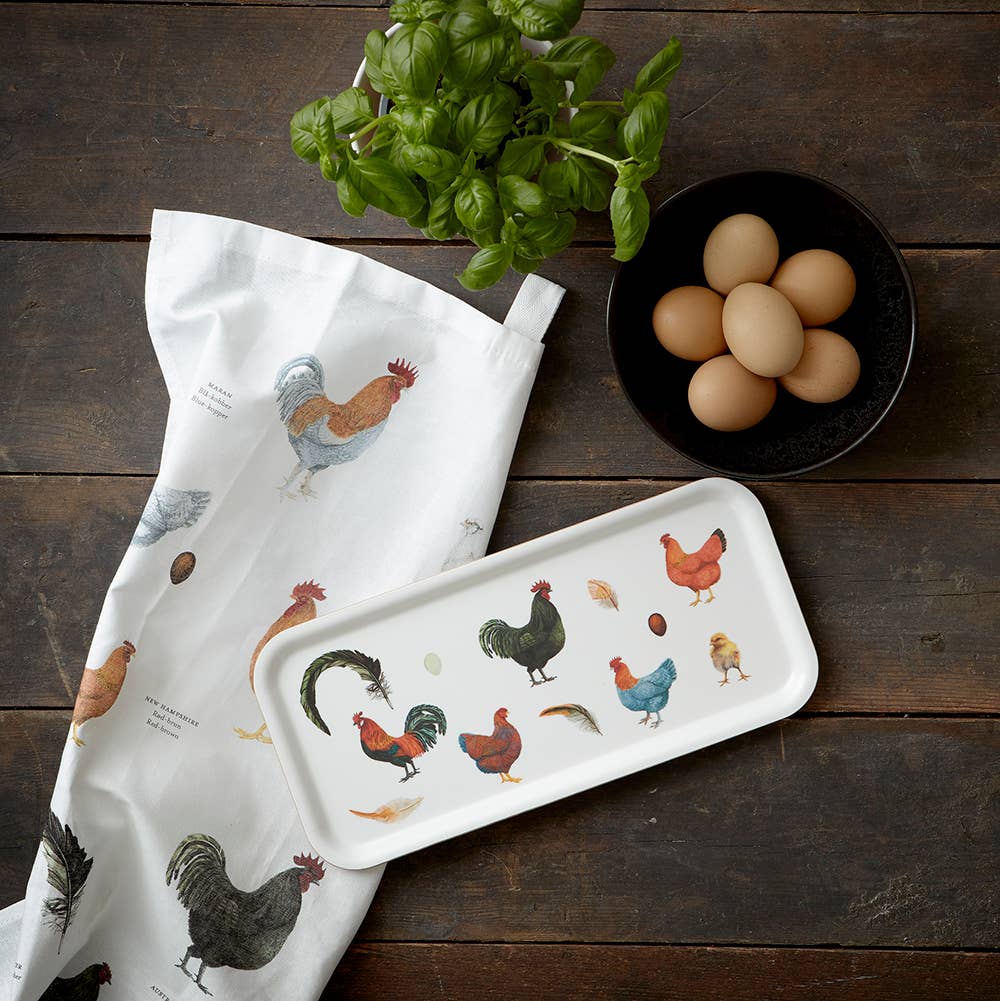 A Colorful Chickens tea towel by Annemarie Haase, a matching rectangular tray, a small plant, and a bowl of brown eggs are elegantly arranged on the wooden surface.