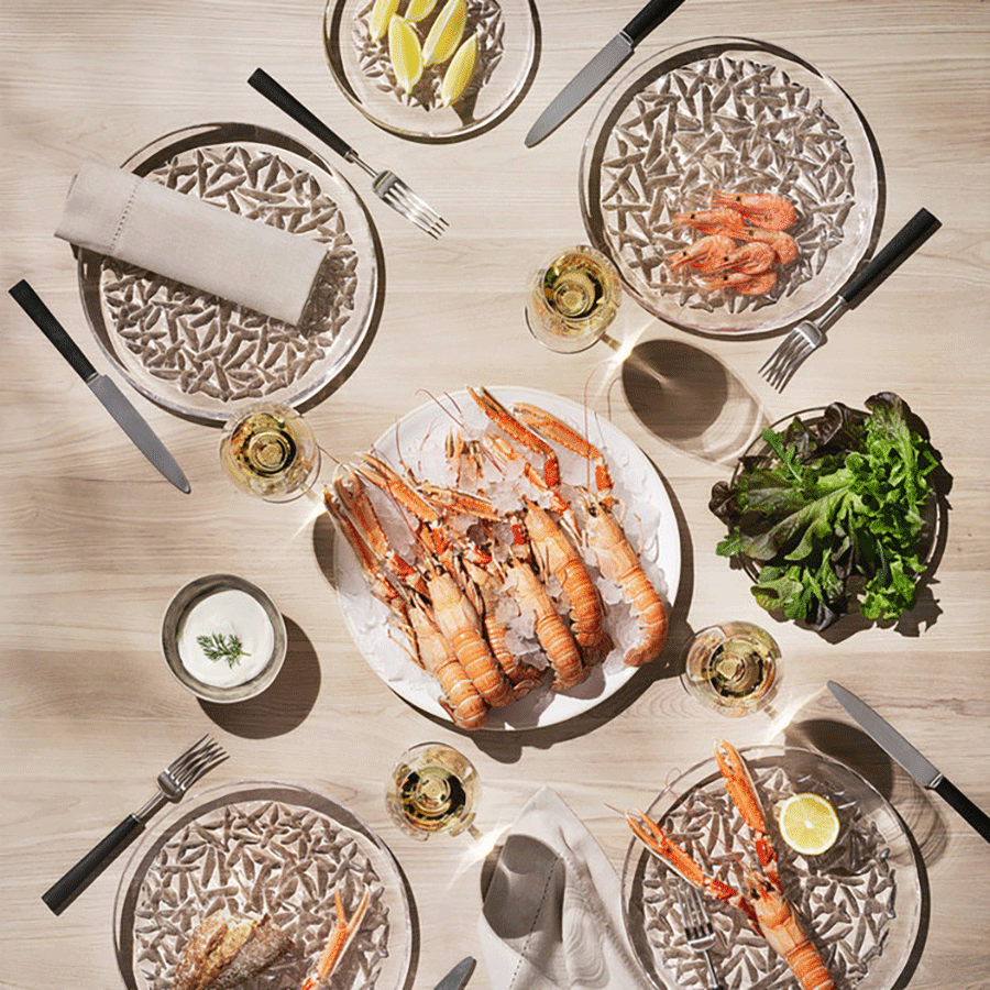 Table set for four with Orrefors: Carat Dinner Plate Designed by Lena Bergstrom, plates of shrimp, langoustines, lemon wedges, mixed greens, sauce, cutlery, and Orrefors crystal wine glasses seen from above.