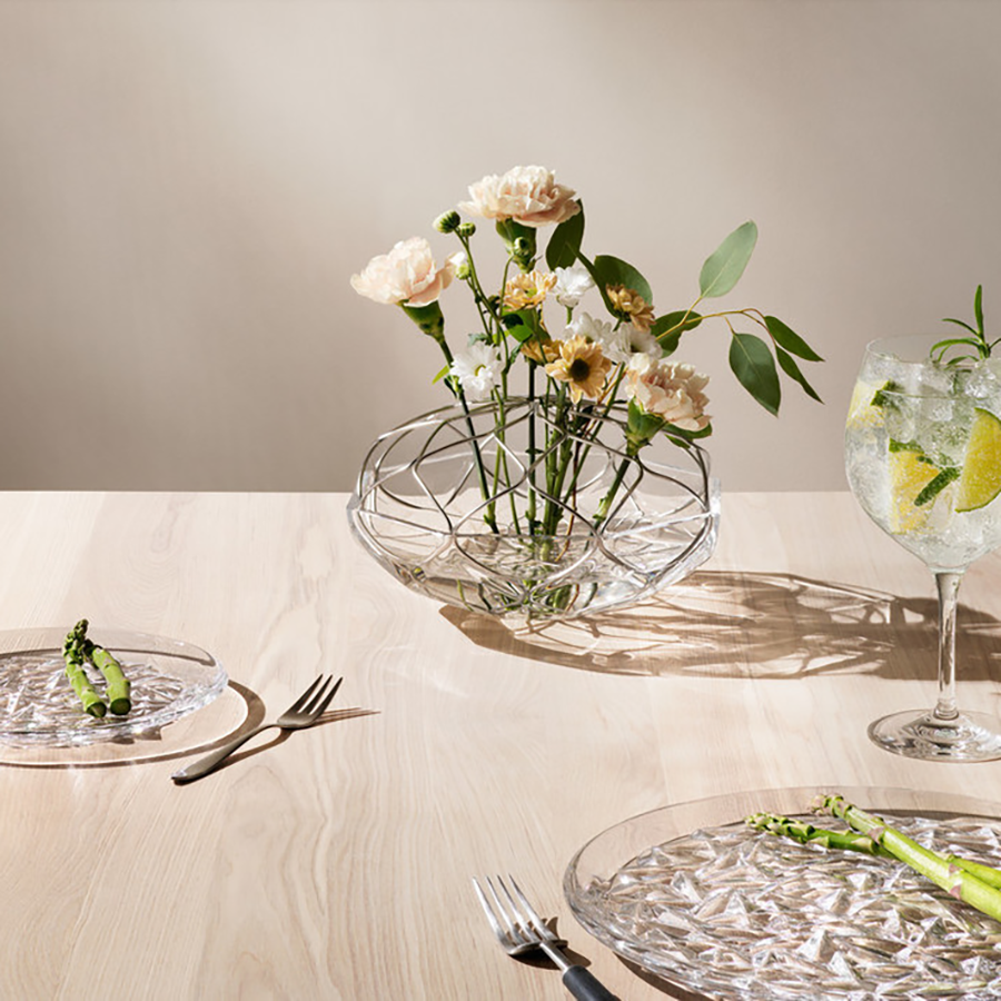 A light wood table showcases asparagus served on Orrefors: Carat Dinner Plate Designed by Lena Bergstrom, accompanied by cutlery, a floral centerpiece in a glass bowl, and a sparkling drink in an Orrefors crystal glass with lime and mint.