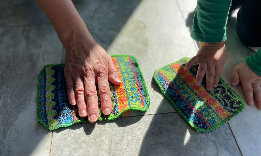 An adult and a child clean a tiled floor with Scrubbie: Skrubba XL Scrub Cloths, enjoying scratch-free scrubbing as sunlight casts shadows on their hands.