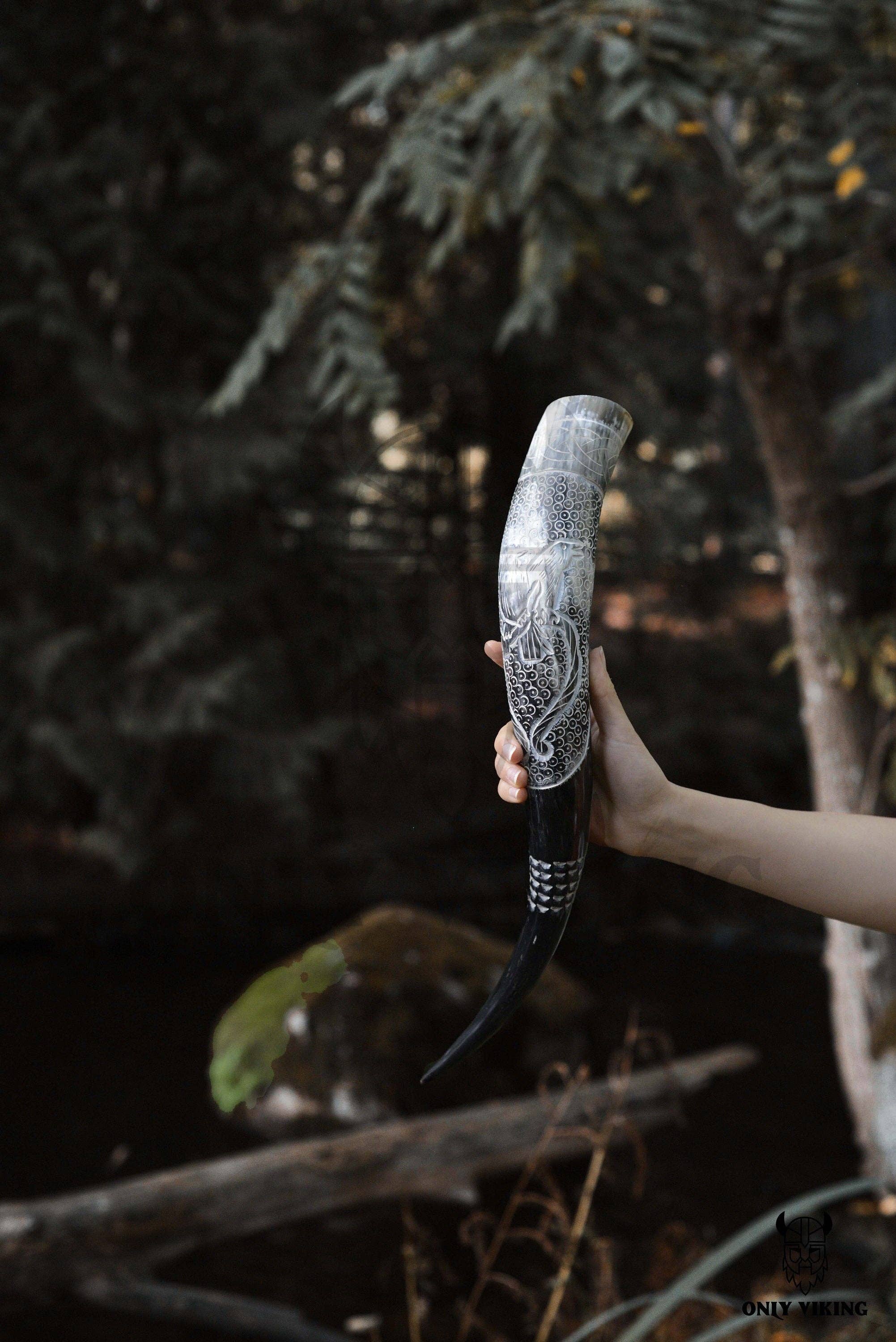 A hand displays the Horn: Raven Viking Drinking Horn w/ Metal Stand (14"-18"), hand-carved with intricate raven designs, set outdoors with green trees and branches in the background.