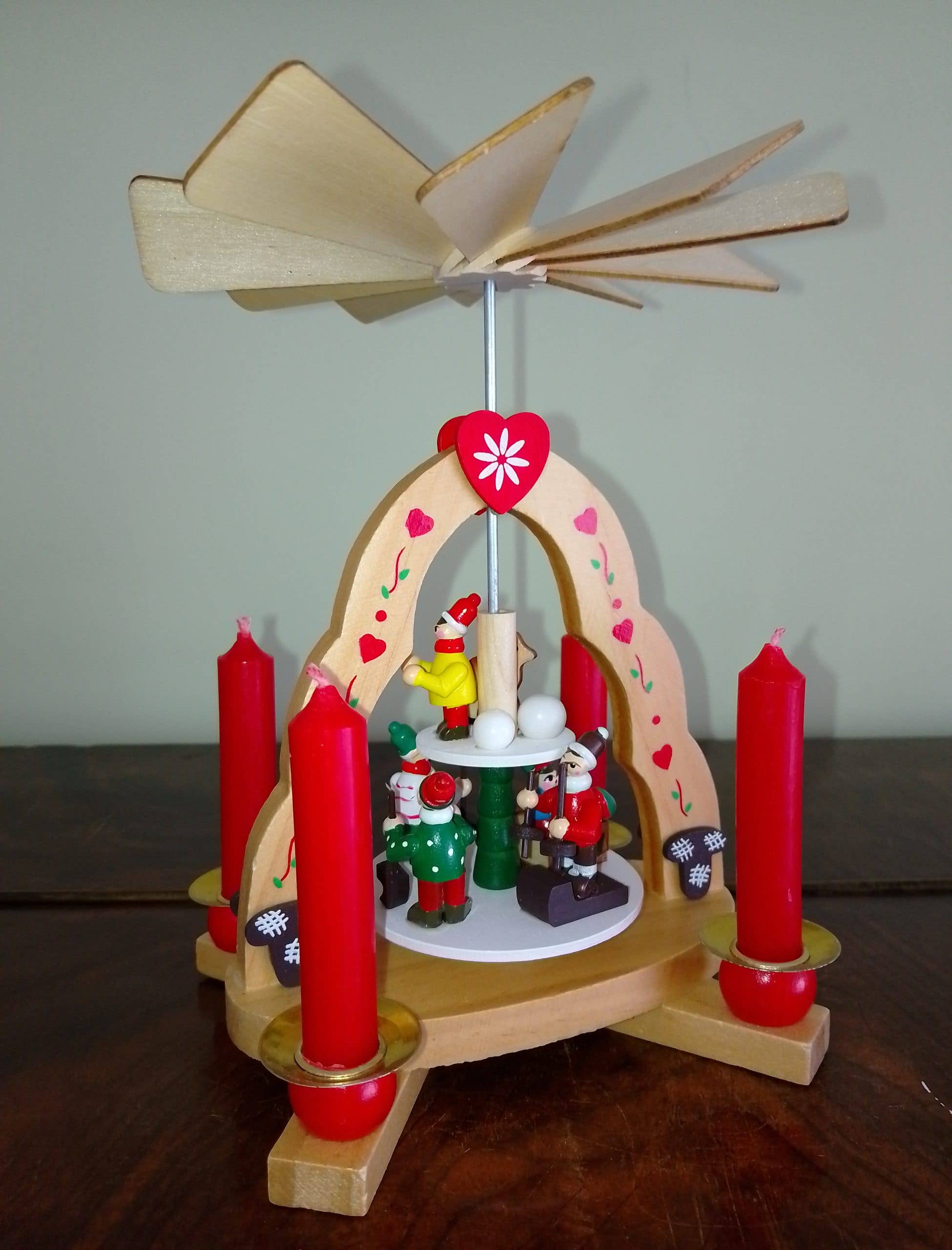 The "Kids" Pyramid Candle Holder, handcrafted in Germany, stands 7" tall by 6" wide, holds four candles, has spinning blades on top, and features small festive figurines at its base.