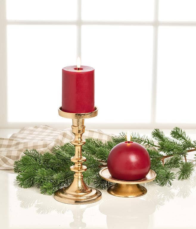 A battery-operated pillar candle in a gold pedestal holder beside a round red LED ball candle on a gold plate, surrounded by evergreen branches.