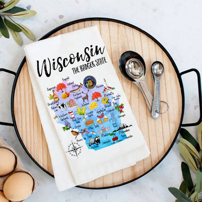 This vibrant Tea Towel, made in the USA, showcases a colorful map of Wisconsin with illustrations of notable landmarks and icons. Labeled The Badger State, it is displayed on a black tray alongside measuring spoons and eggs.