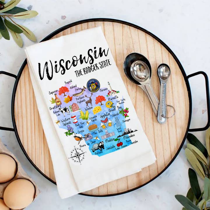 This vibrant Tea Towel, made in the USA, showcases a colorful map of Wisconsin with illustrations of notable landmarks and icons. Labeled The Badger State, it is displayed on a black tray alongside measuring spoons and eggs.