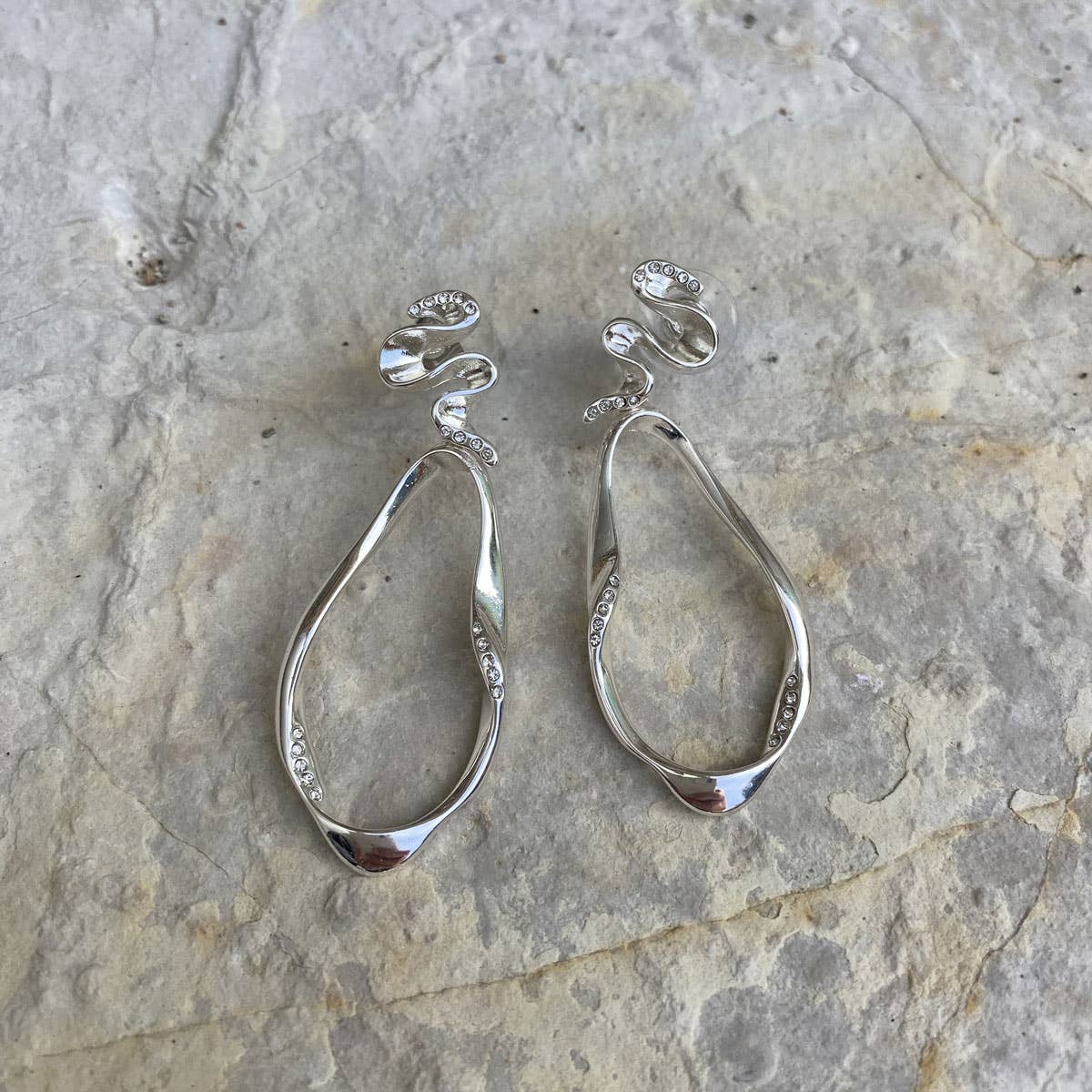 A pair of Blowing Ribbon Silver Dangle Earrings by A&C Oslo, featuring an irregular ribbon-like shape with small crystals, displayed on a textured stone surface.