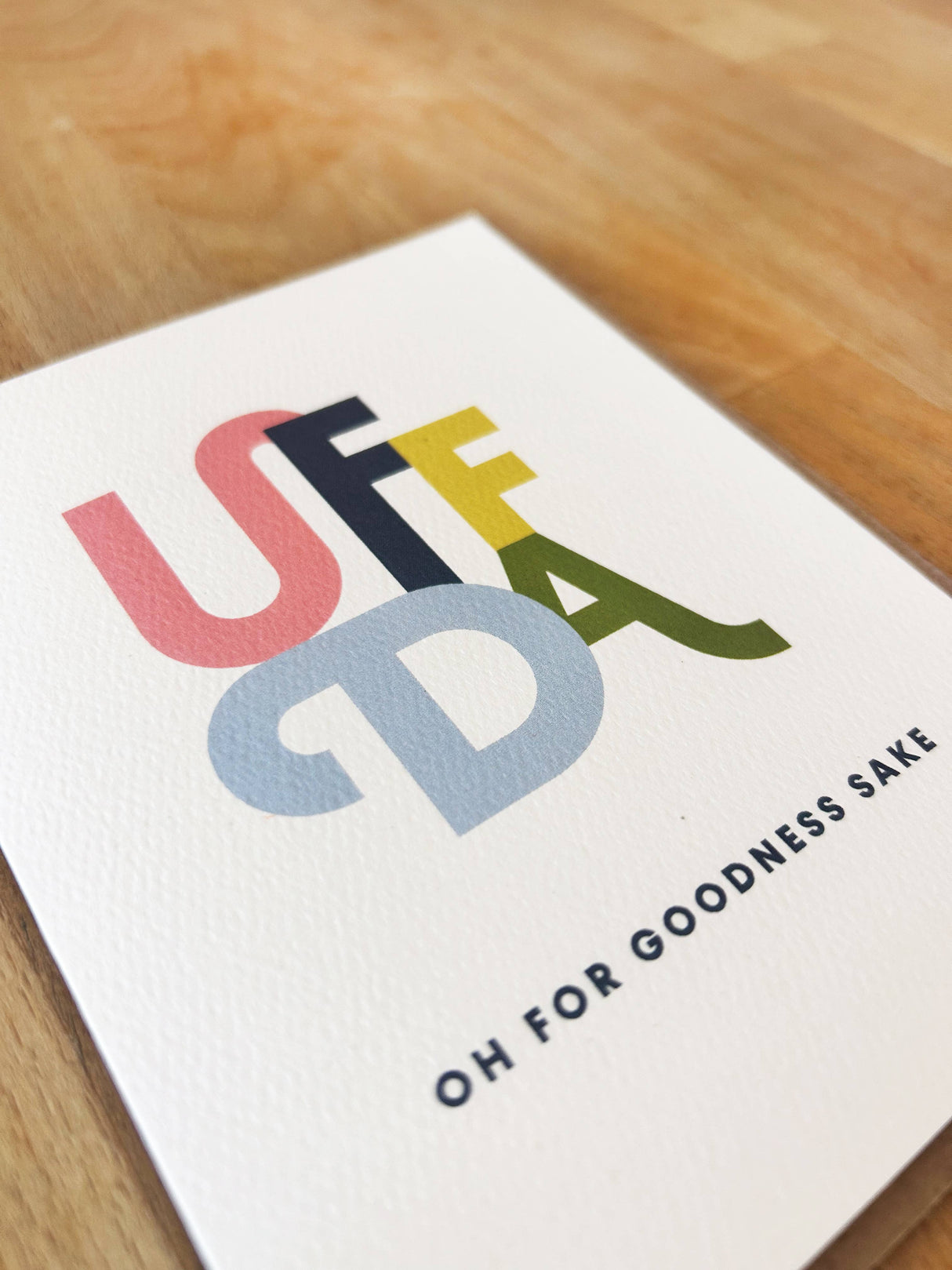 The Uffda Greeting Card (4.25"x5.5", blank inside) features bold, colorful "UFF DA" letters and "OH FOR GOODNESS SAKE" in black text on the front, shown resting on wood; includes a brown envelope.