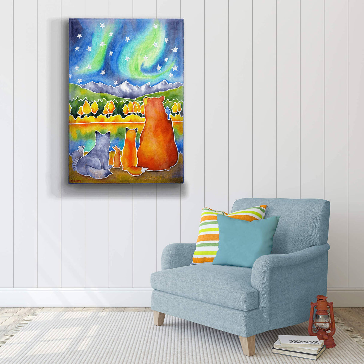 A blue armchair with colorful pillows sits by a wall decorated with "Drawn to the Lights" metal box art (18" x 26"), depicting Scandinavian woodland animals under northern lights.