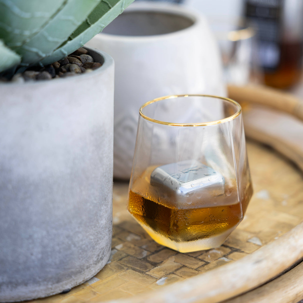 A glass of whiskey with Cubes: Stainless Steel Ice Cubes in Plastic Case Set of 2, 1.57" Square sits on a tray next to two potted plants in concrete planters.