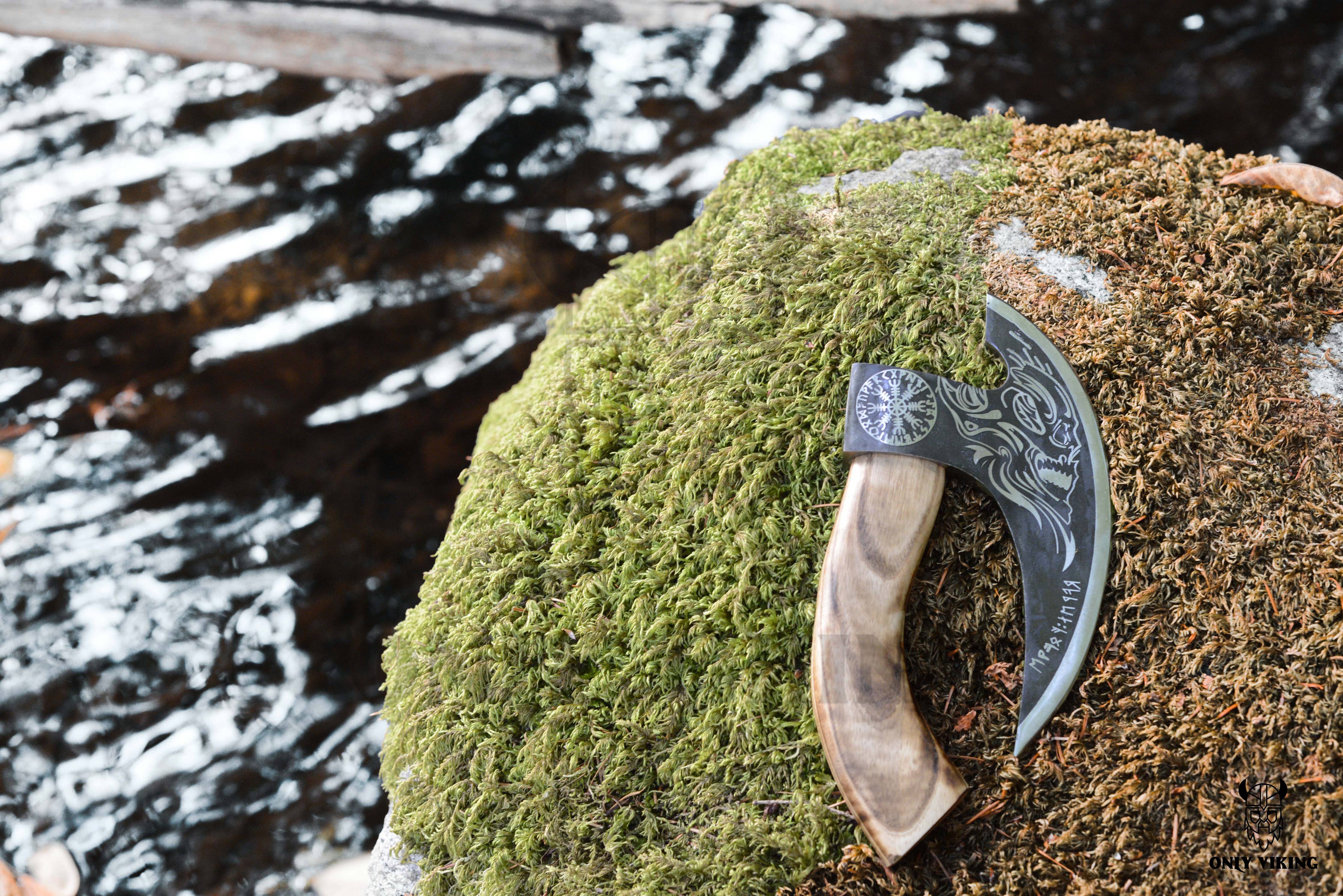A pizza cutter with an axe design and leather sheath rests on a moss-covered rock beside flowing water, its stainless steel blade glinting softly in the dappled sunlight.